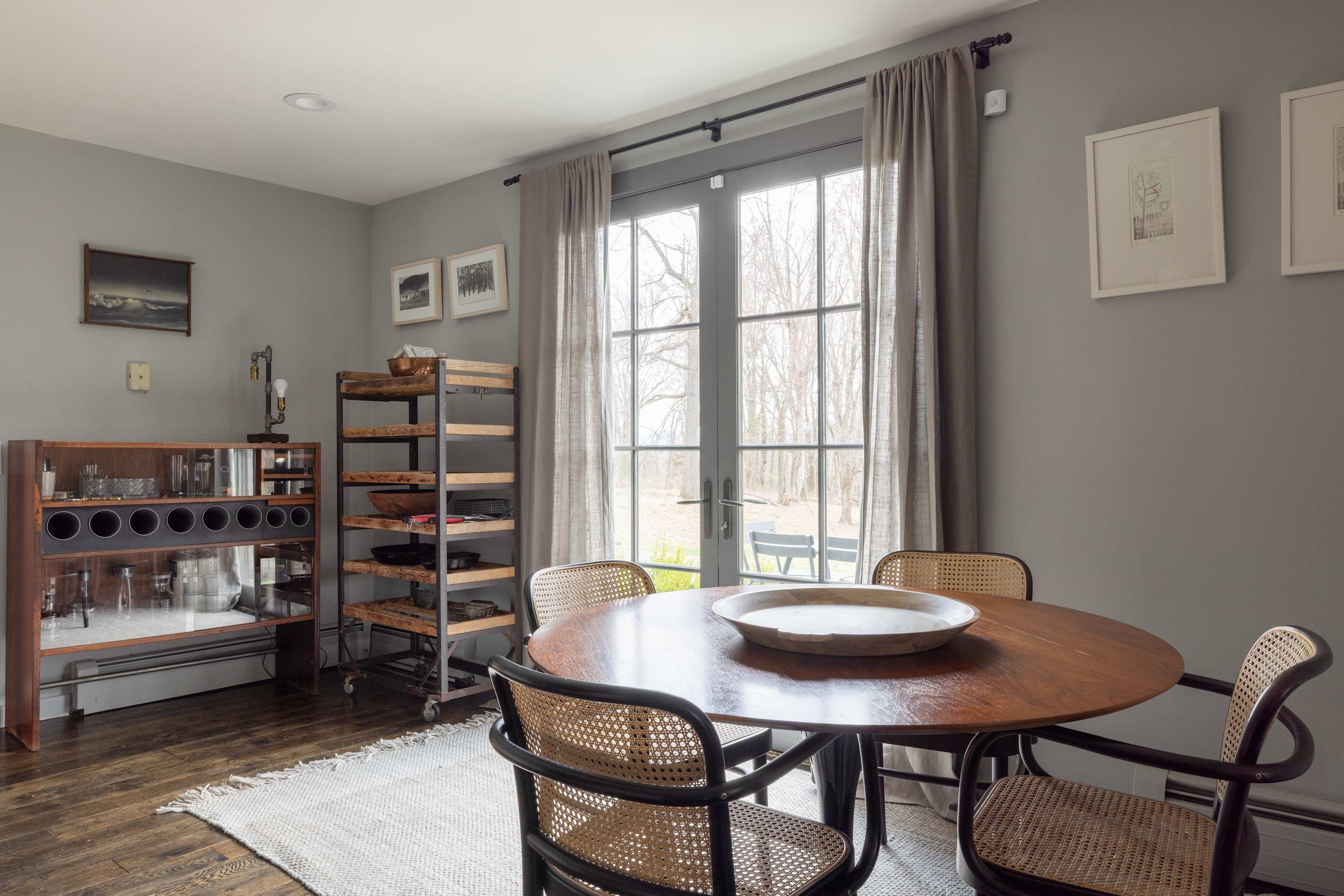 Dining area with a round wooden table and four cane-back chairs, set on a light woven rug. French doors lead to an outdoor space, flanked by sheer brown curtains. A wooden bar cart and industrial shelving unit are also visible.