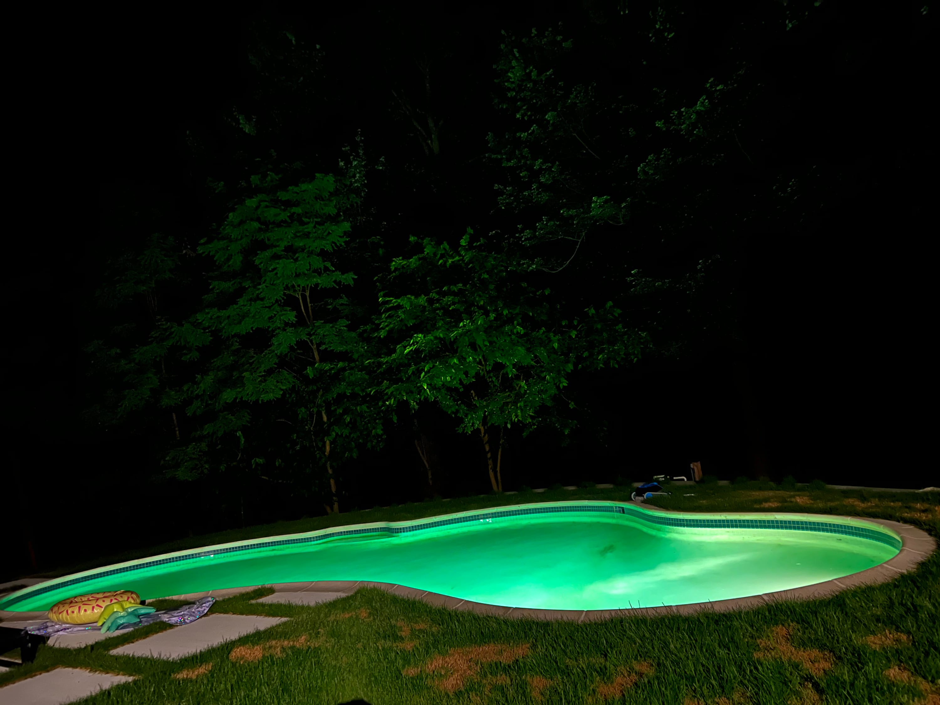 The swimming pool glows with vibrant green underwater lighting at night, surrounded by dark grass and silhouetted trees. A yellow inflatable donut float is visible on the pool deck.