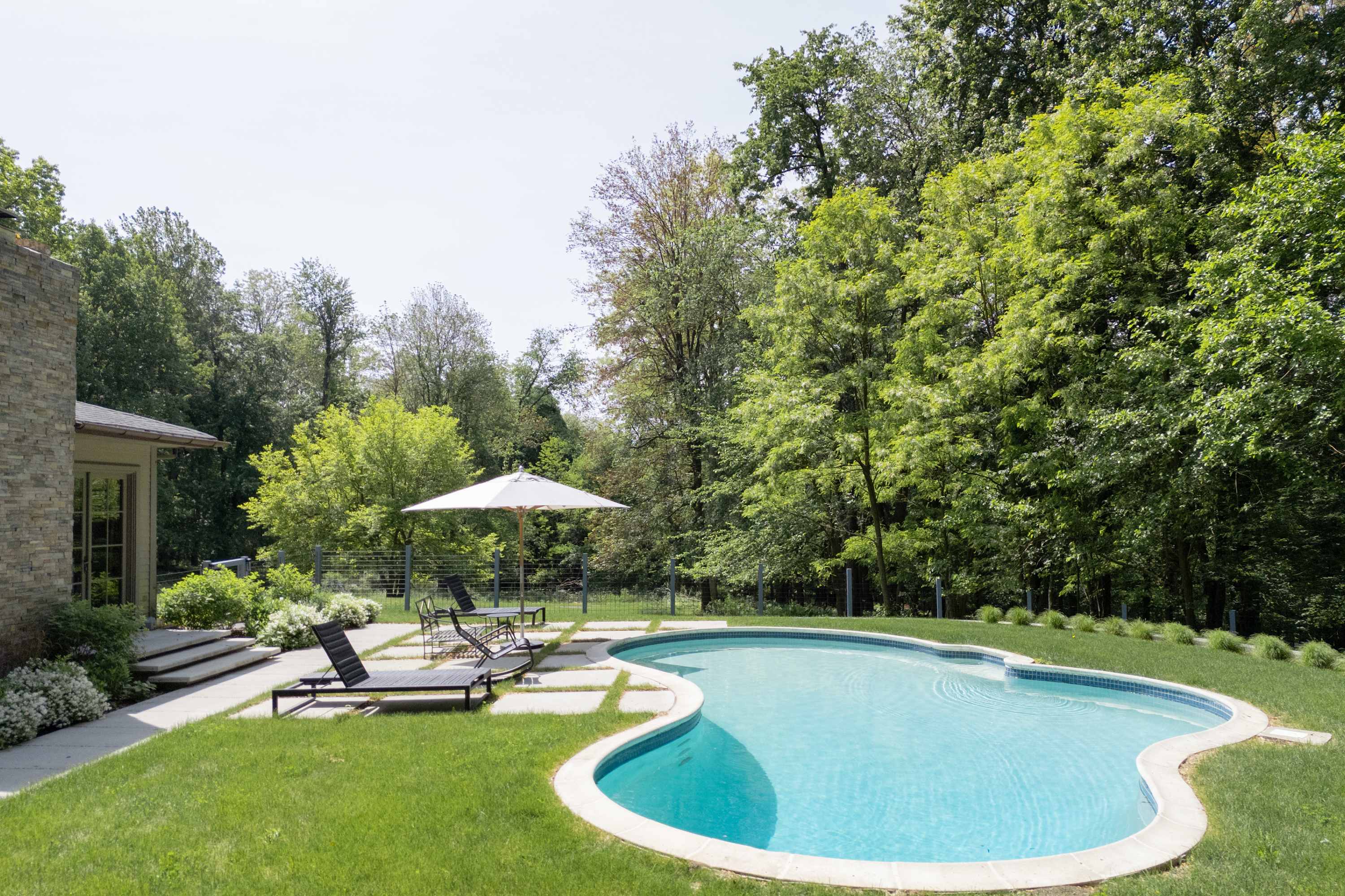 A serene swimming pool with a light blue interior, surrounded by a well-maintained lawn and a patio with square stepping stones. Black lounge chairs and a white umbrella are arranged for relaxation, with a dense forest providing a natural backdrop.