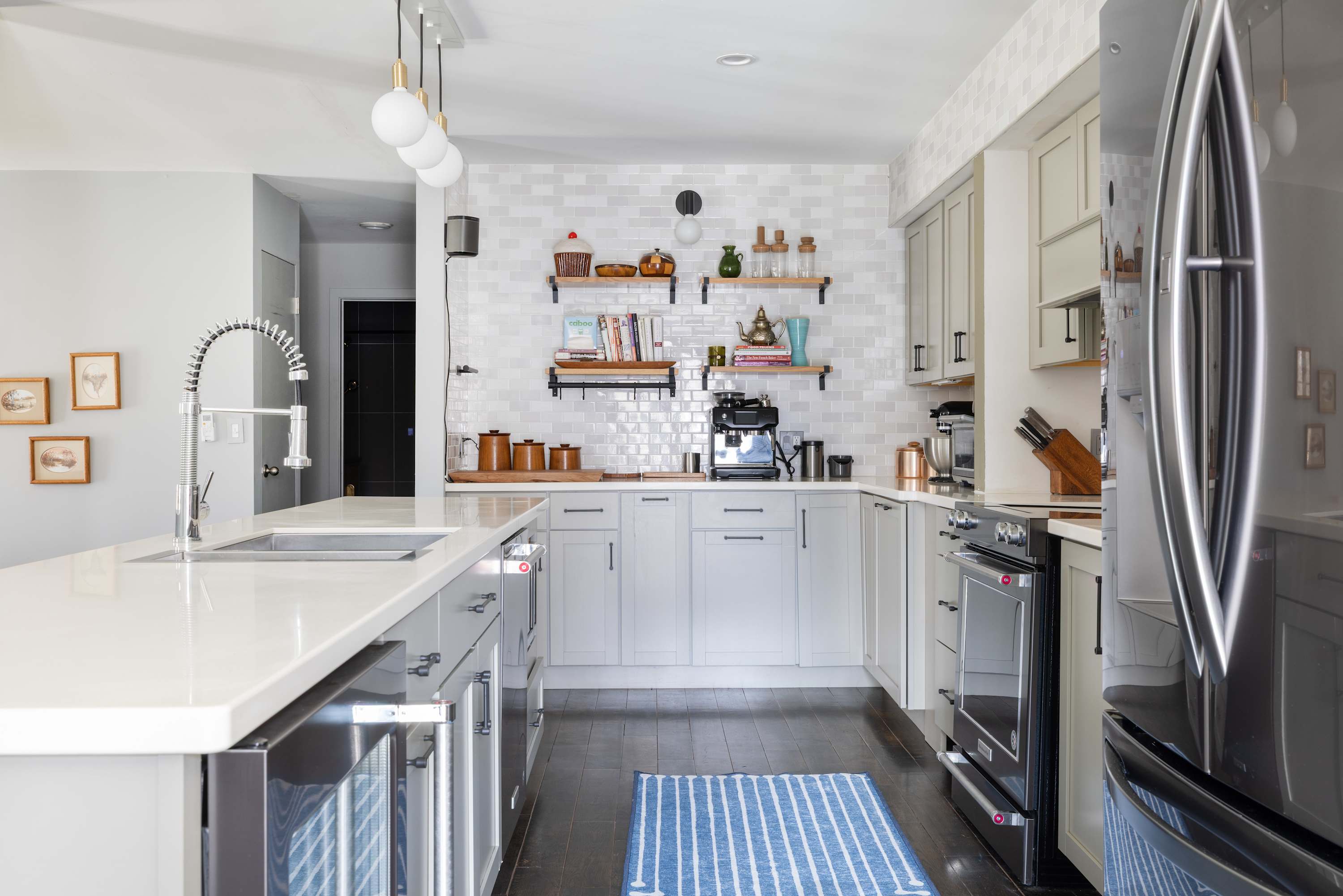 A bright kitchen with light gray cabinets and white countertops features a large island with an integrated sink and gooseneck faucet, a black range, and open shelving against a white tiled wall, with dark wood floors and a blue striped rug.