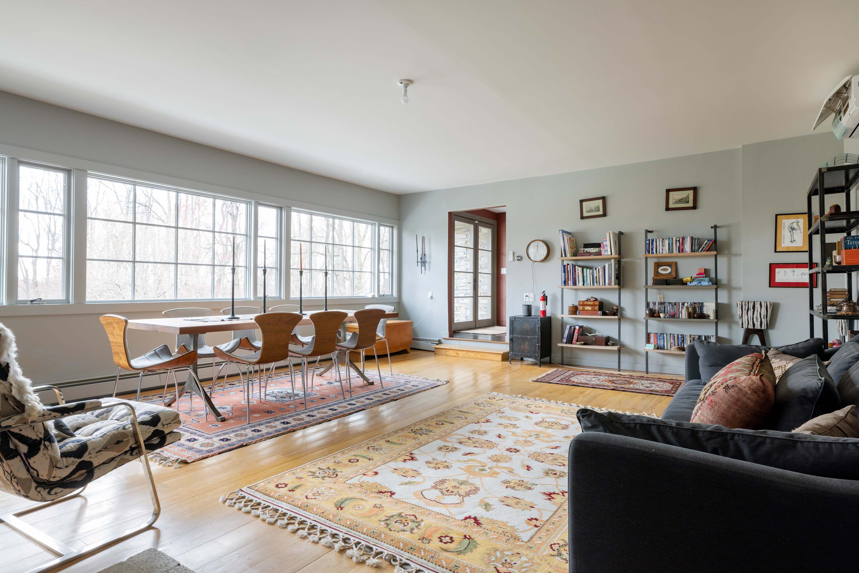 A large open-concept living and dining area features light wood floors, a long dining table with modern chairs by a wall of windows, and a dark gray sofa with bookshelves, all illuminated by natural light.