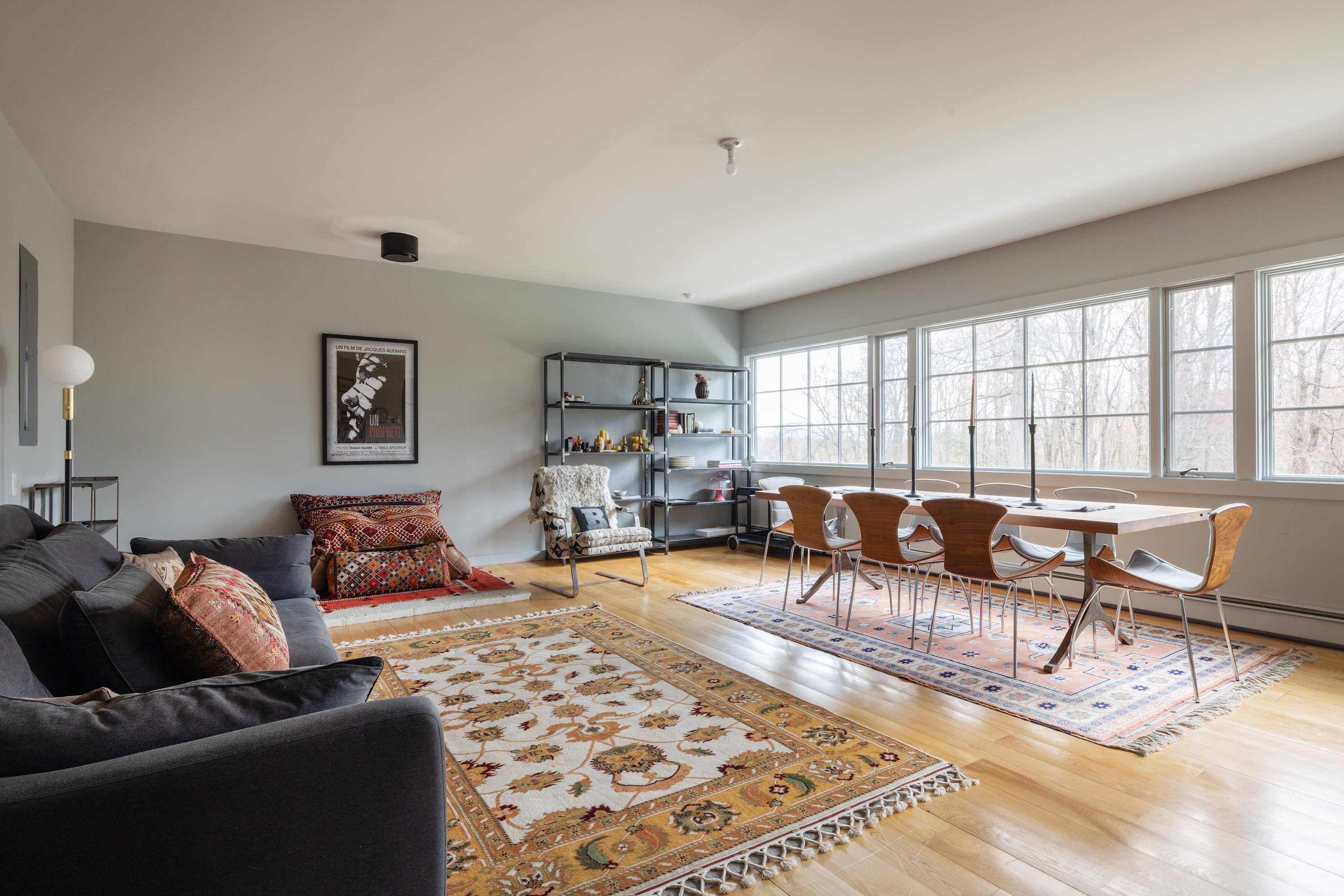 Expansive open-concept living and dining area with light wood floors and large windows offering natural light. A gray sofa with patterned pillows and a long dining table with modern chairs are arranged on colorful rugs.
