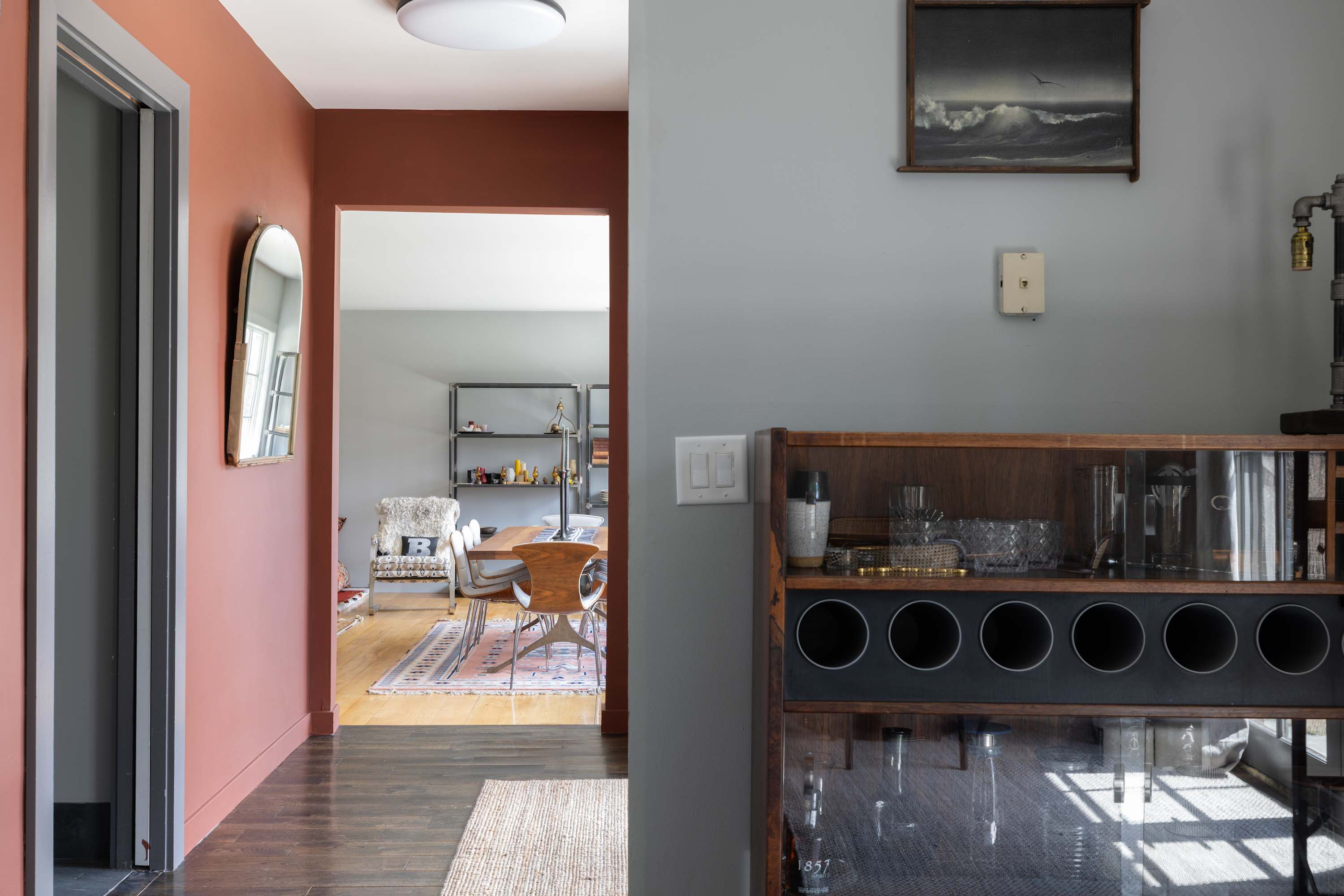 Hallway with a warm terracotta accent wall and a mirror, leading into an open-concept living/dining area with light wood floors. A dark wood bar cabinet with glass storage is visible in the foreground.