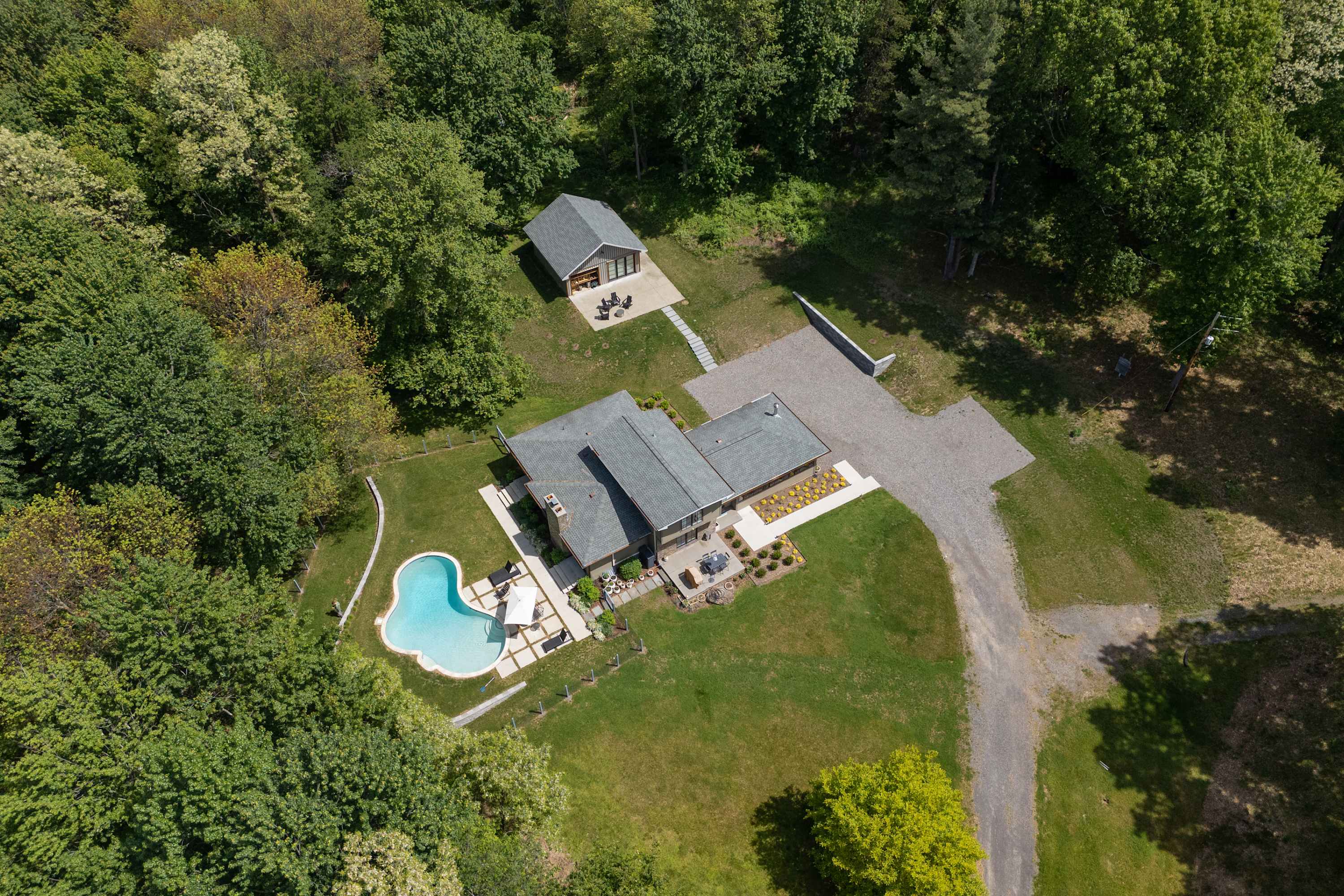 An aerial view showcasing the main house and a separate utility building nestled within a lush green landscape of trees. A uniquely shaped swimming pool is visible next to the main house, connected by a gravel driveway.