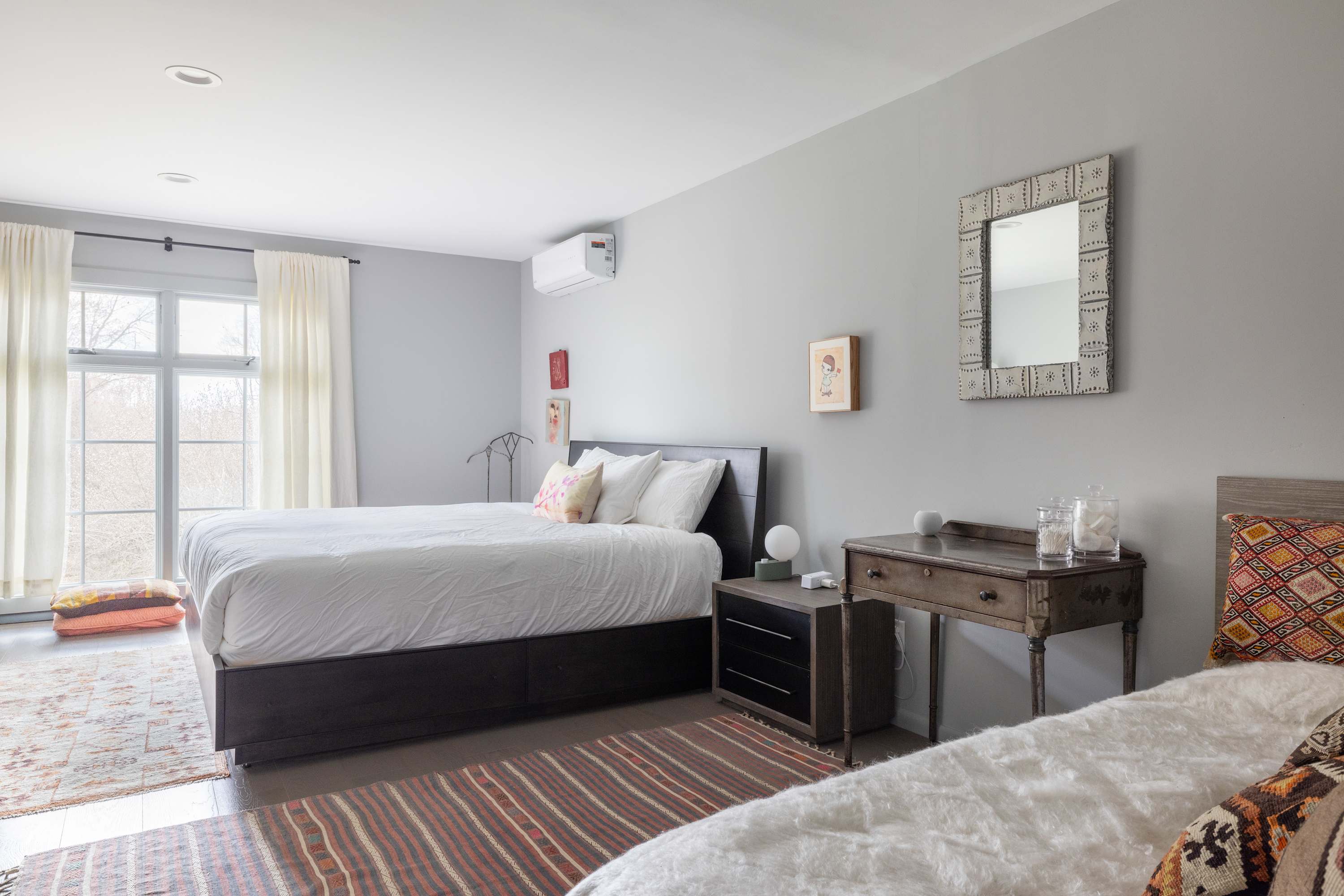 A bright bedroom with light gray walls, a large bed with white bedding, and a smaller bed partially visible on the right. A decorative mirror hangs above a wooden desk, and large windows with sheer curtains offer natural light.