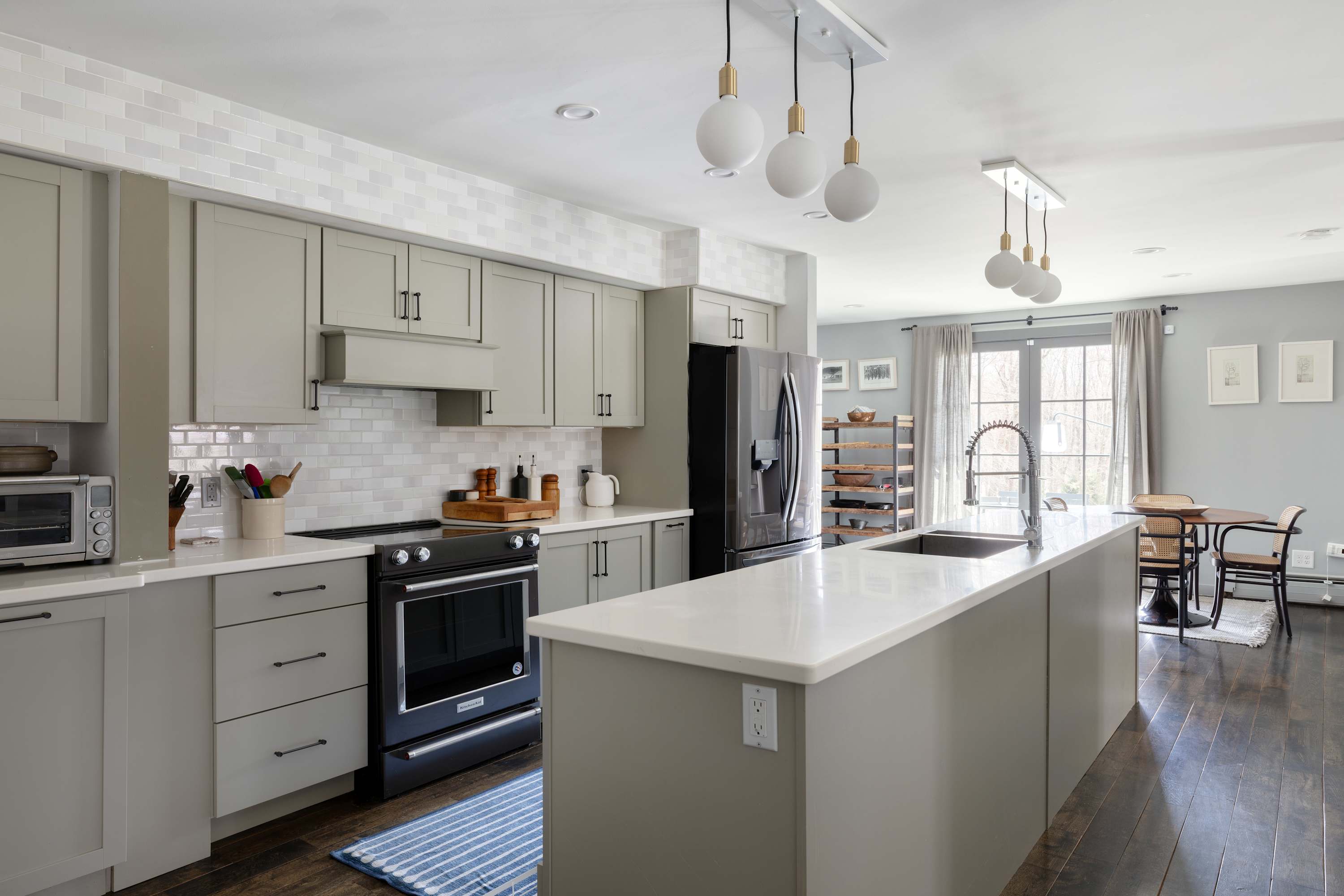 A modern kitchen with light gray shaker cabinets, white countertops, and a white subway tile backsplash features a large island with a sink, a black range, and stainless steel appliances, with dark wood floors extending into an adjacent dining area.