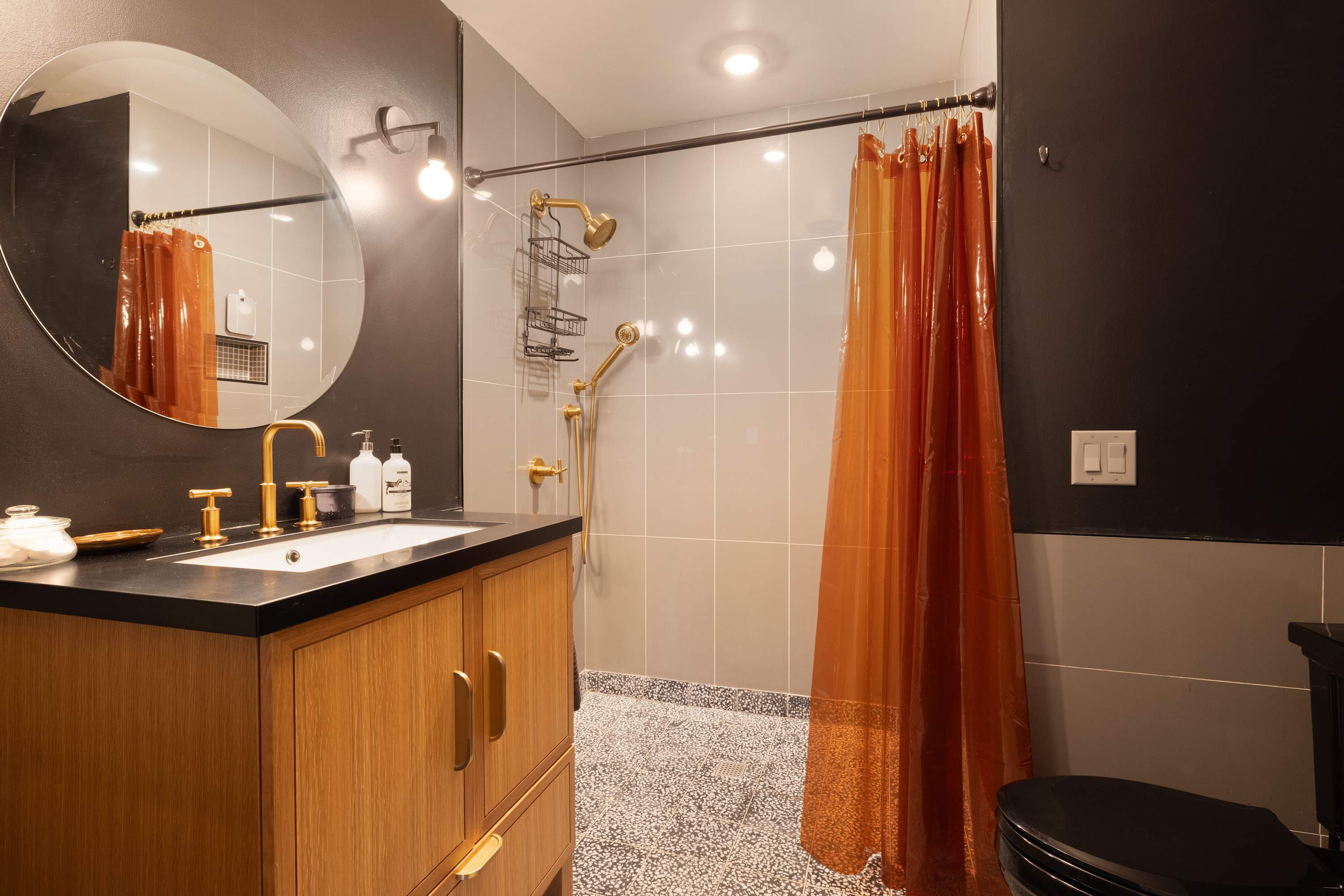 Modern bathroom with a wooden vanity, black countertop, and gold fixtures, including a round mirror. The shower features light gray subway tiles, a gold showerhead, and a vibrant orange shower curtain.