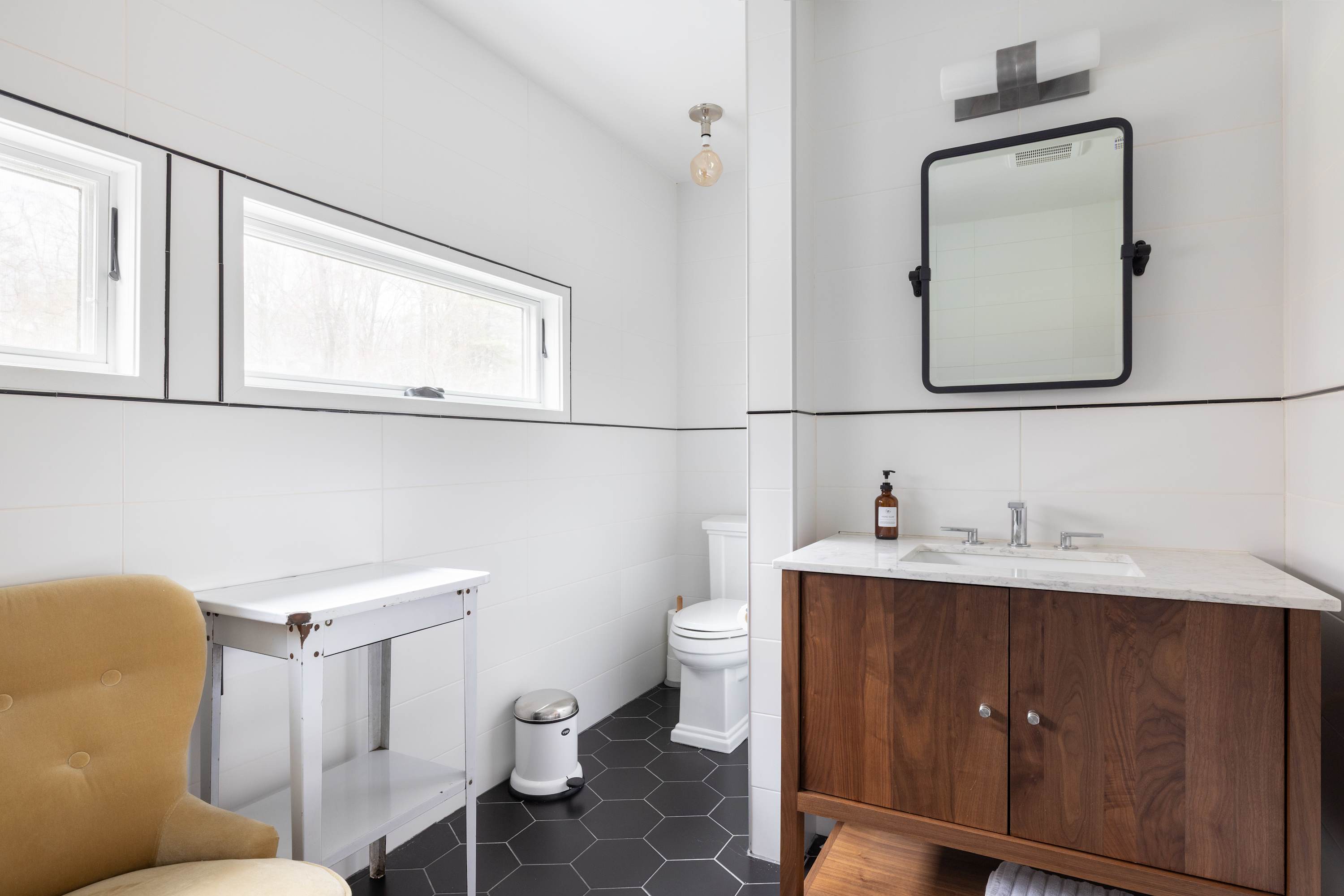 A modern bathroom with white subway tiles and black accent lines, featuring a dark wood vanity with a white marble top and a rectangular mirror. Black hexagonal floor tiles and two horizontal windows complete the bright space.