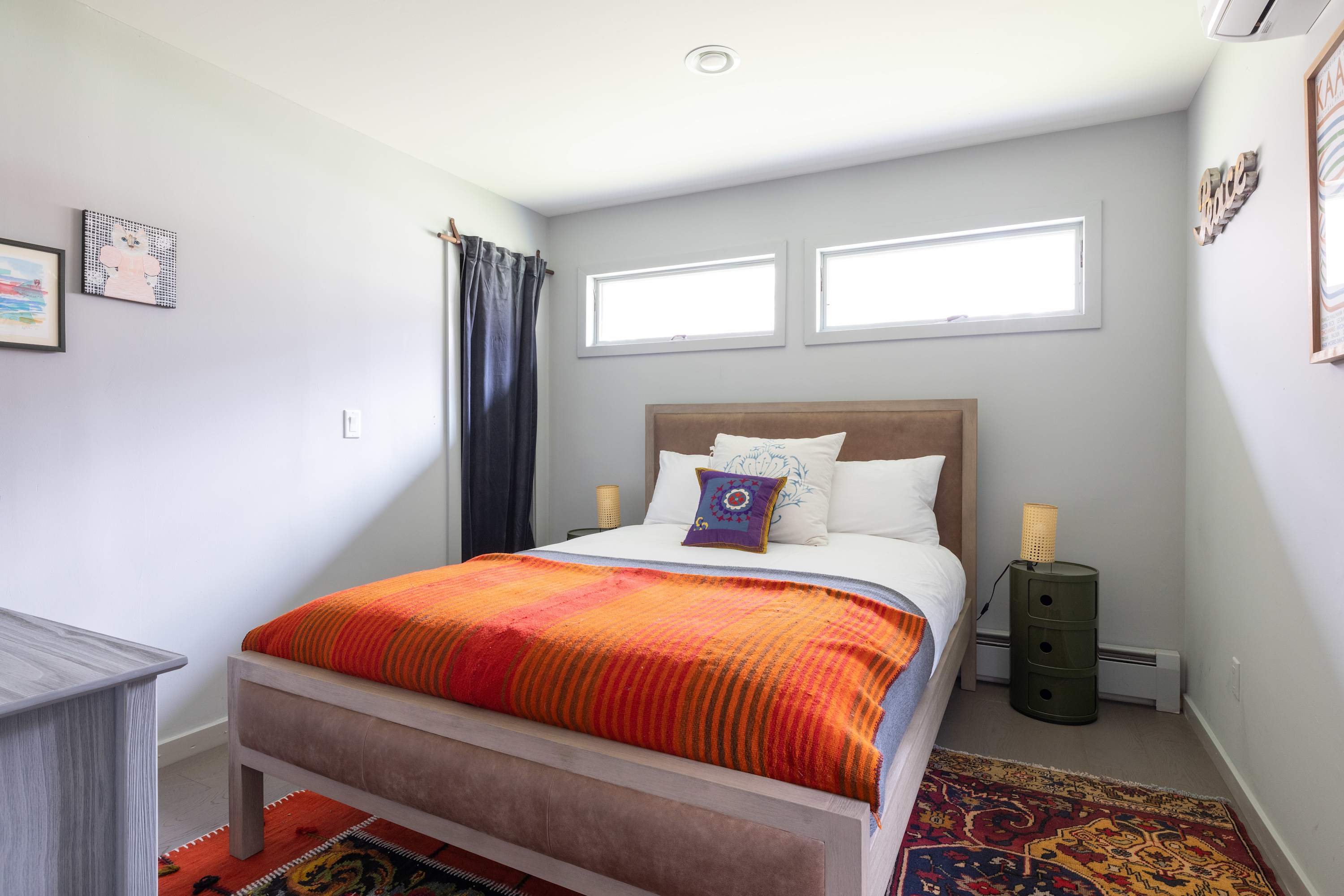 Serene bedroom with a queen-sized bed featuring a light brown upholstered headboard and an orange and red striped blanket. Two small windows are positioned above the bed, flanked by modern nightstands with woven lamps.