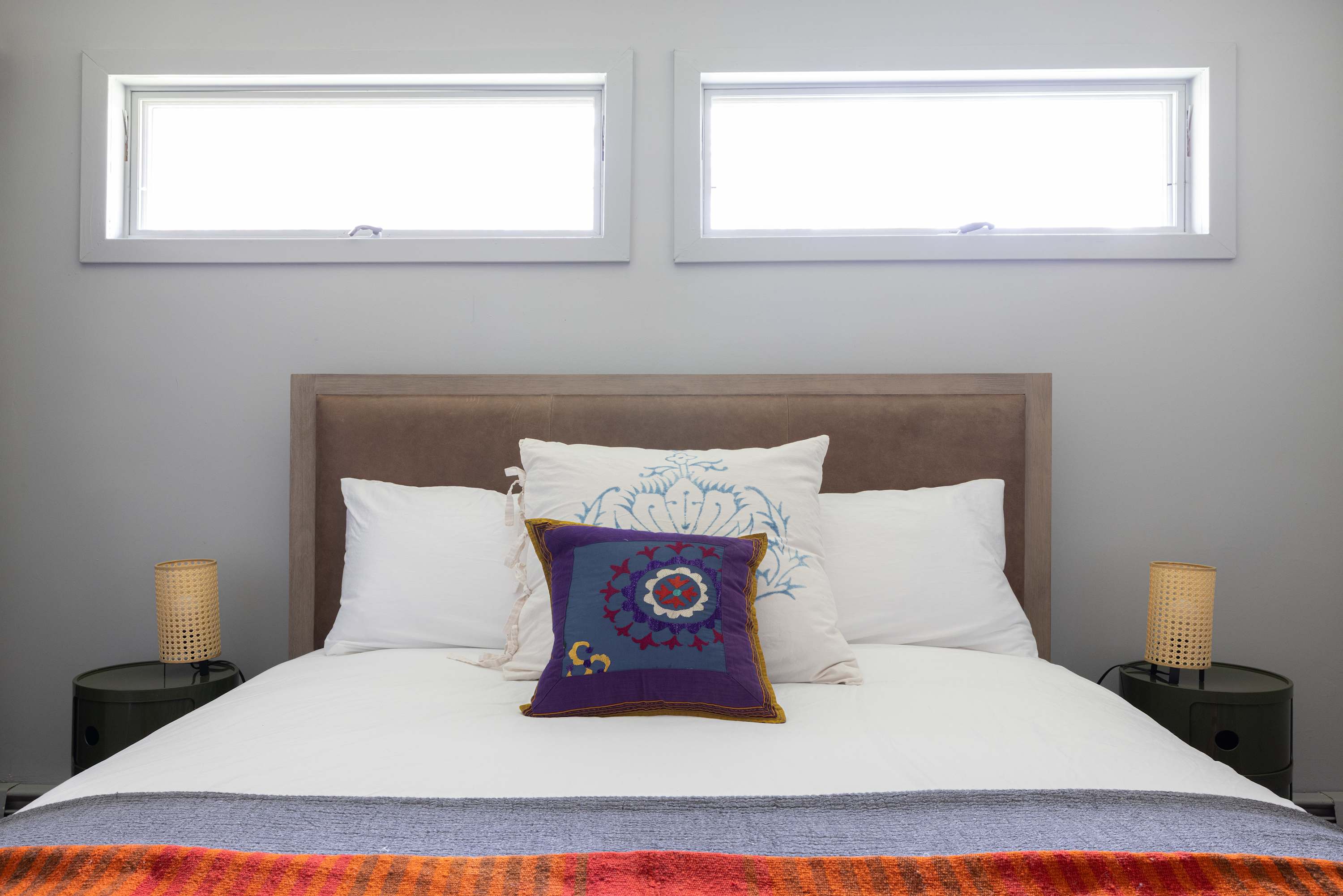 Close-up of a queen bed with a light brown upholstered headboard, white bedding, and a vibrant purple decorative pillow. Two small horizontal windows are centered above the bed, with woven lamps on dark green nightstands.