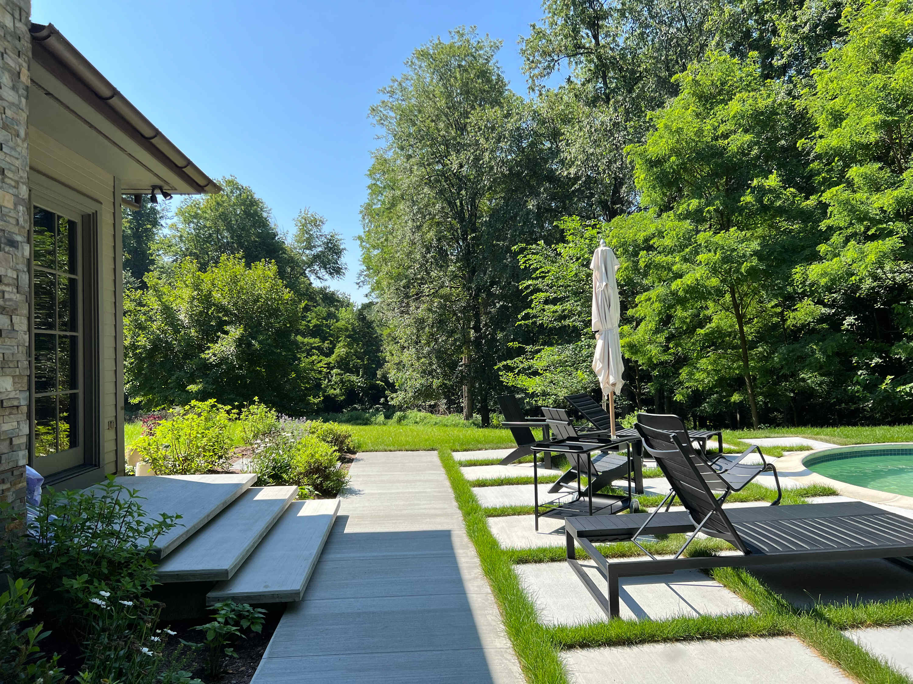 A modern outdoor space featuring a swimming pool with a unique shape, surrounded by a patio with large square pavers and grass inserts. Several black lounge chairs and an umbrella provide relaxation areas, all set against a lush green backdrop of trees.