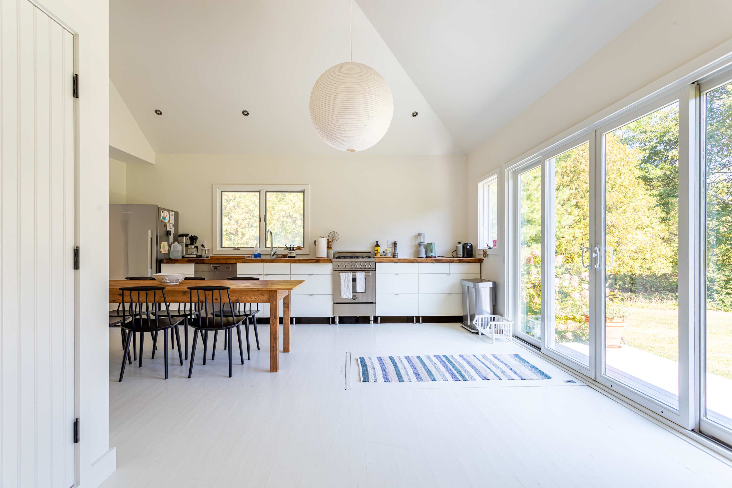 A modern kitchen and dining area with white vaulted ceilings and floors, featuring a long wooden dining table and black chairs. White cabinets and wooden countertops line one wall, with large sliding glass doors opening to the outdoors.