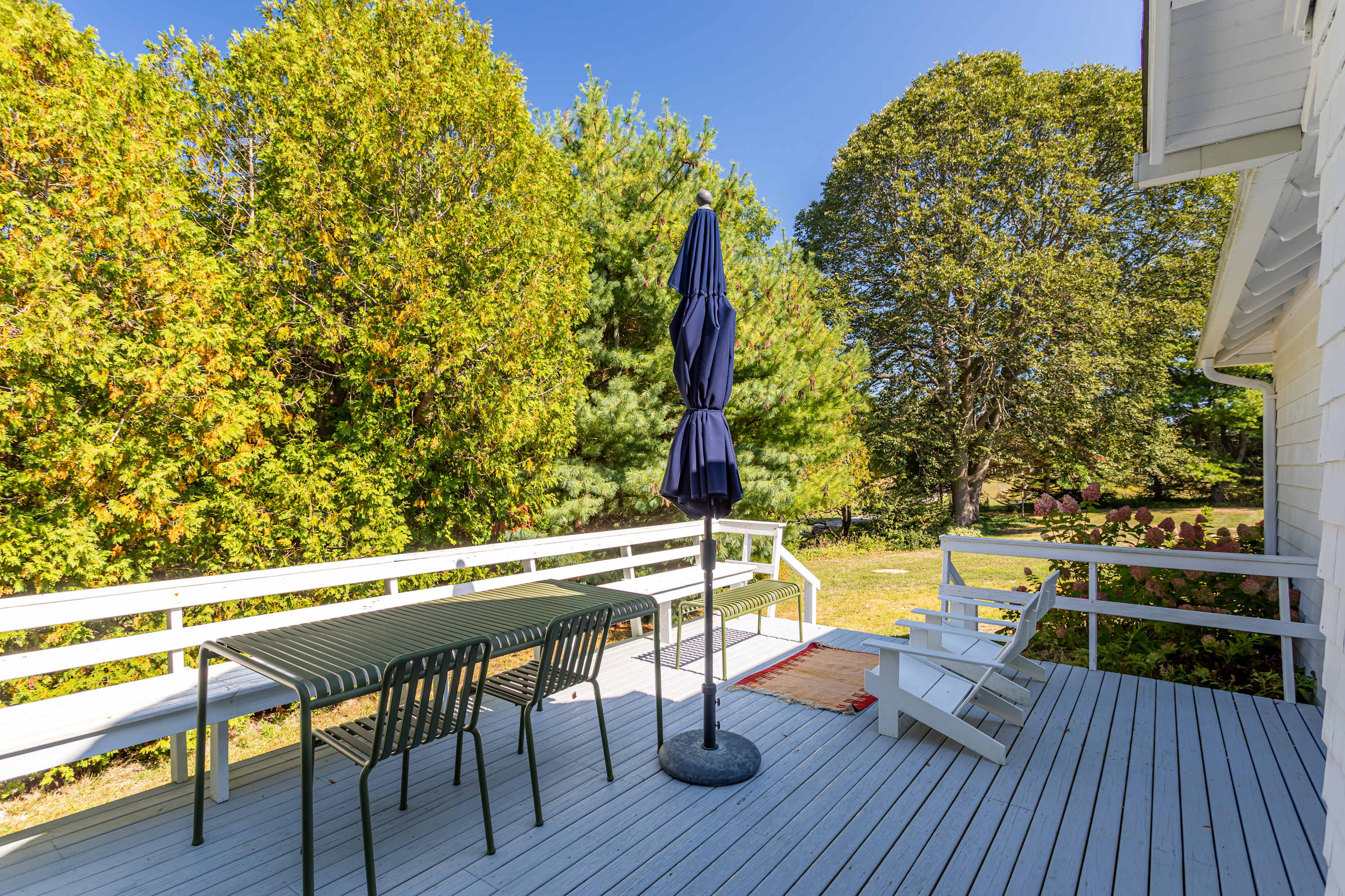 A spacious outdoor deck with light gray wooden planks, featuring a dark green slatted dining table with matching chairs and bench, a closed blue umbrella, and two white Adirondack chairs, all surrounded by lush green trees.