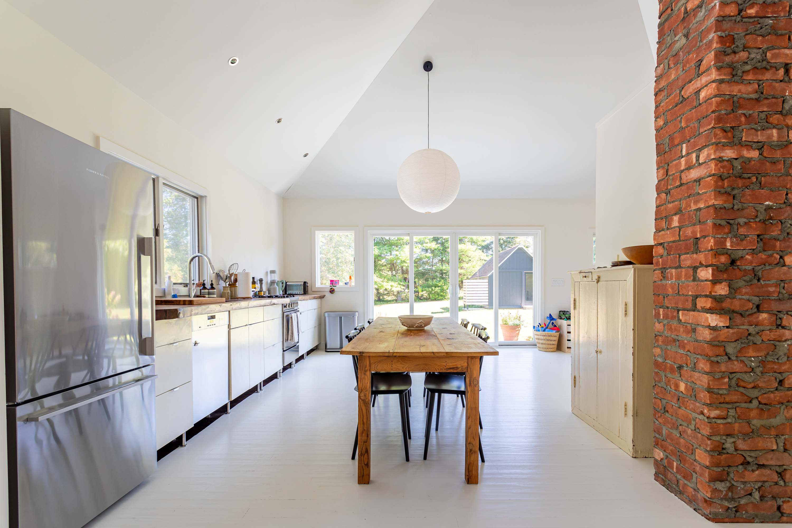 A spacious, open-concept kitchen and dining area with a long wooden dining table and black chairs. The kitchen features white cabinetry, wooden countertops, and stainless steel appliances, with a large brick column adding a rustic touch.