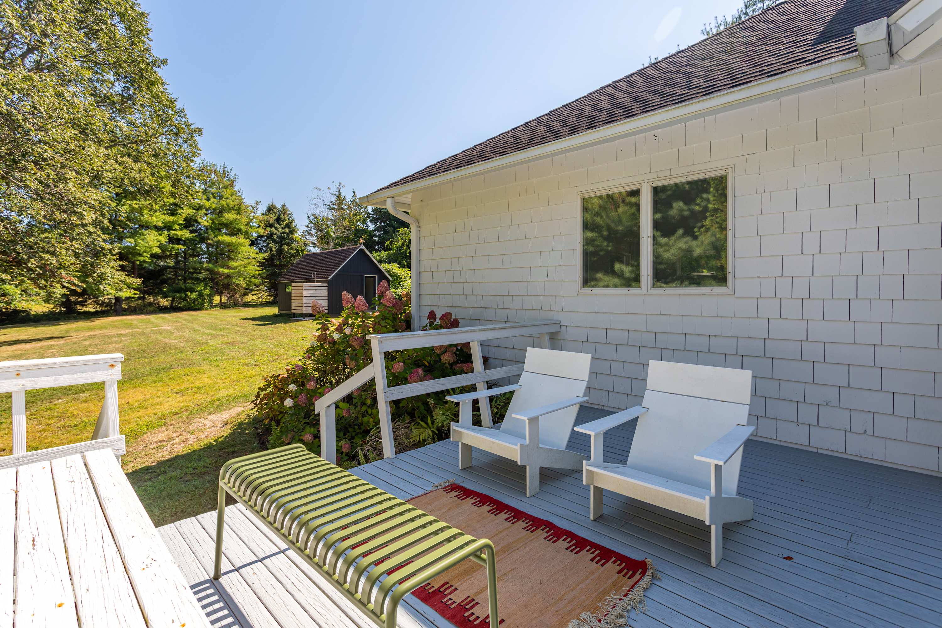 Outdoor deck with grey flooring, featuring two white Adirondack chairs, a green slatted bench, and a colorful rug, with the main house and a distant shed visible.