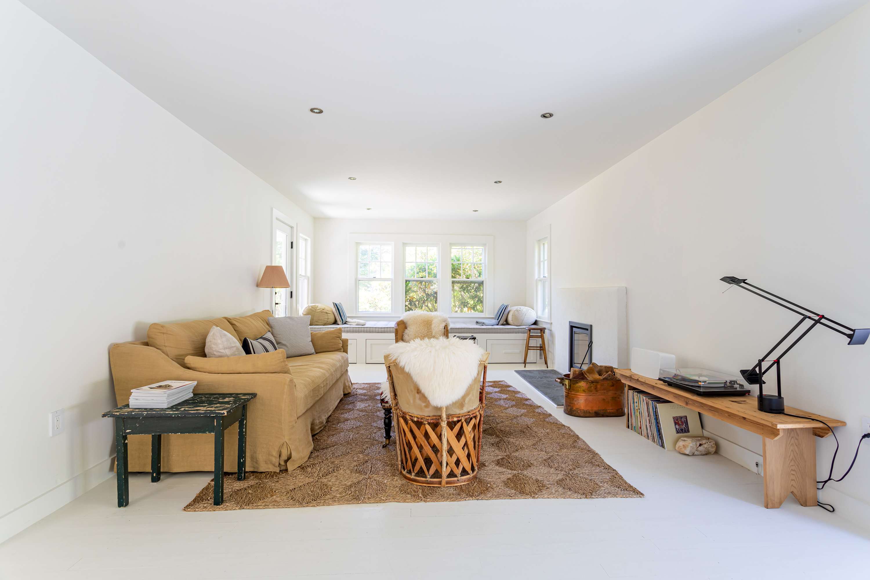 Spacious living room with crisp white walls and floors, a mustard yellow sofa, a textured patterned rug, a long window seat, and a modern fireplace, creating a bright and inviting atmosphere.