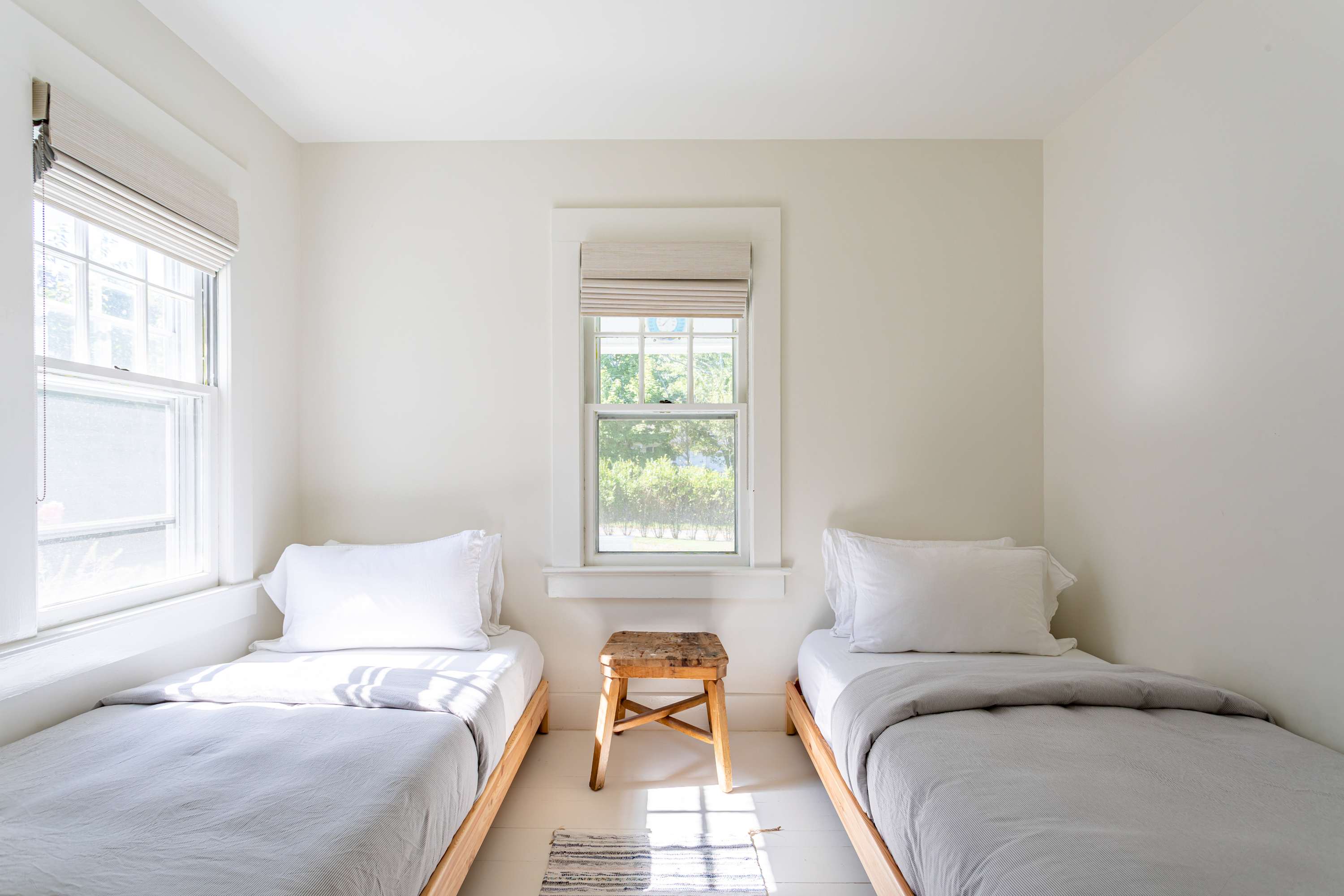 Bright bedroom with two twin beds featuring light grey bedding and wooden frames, separated by a rustic wooden stool, and two windows with blinds.