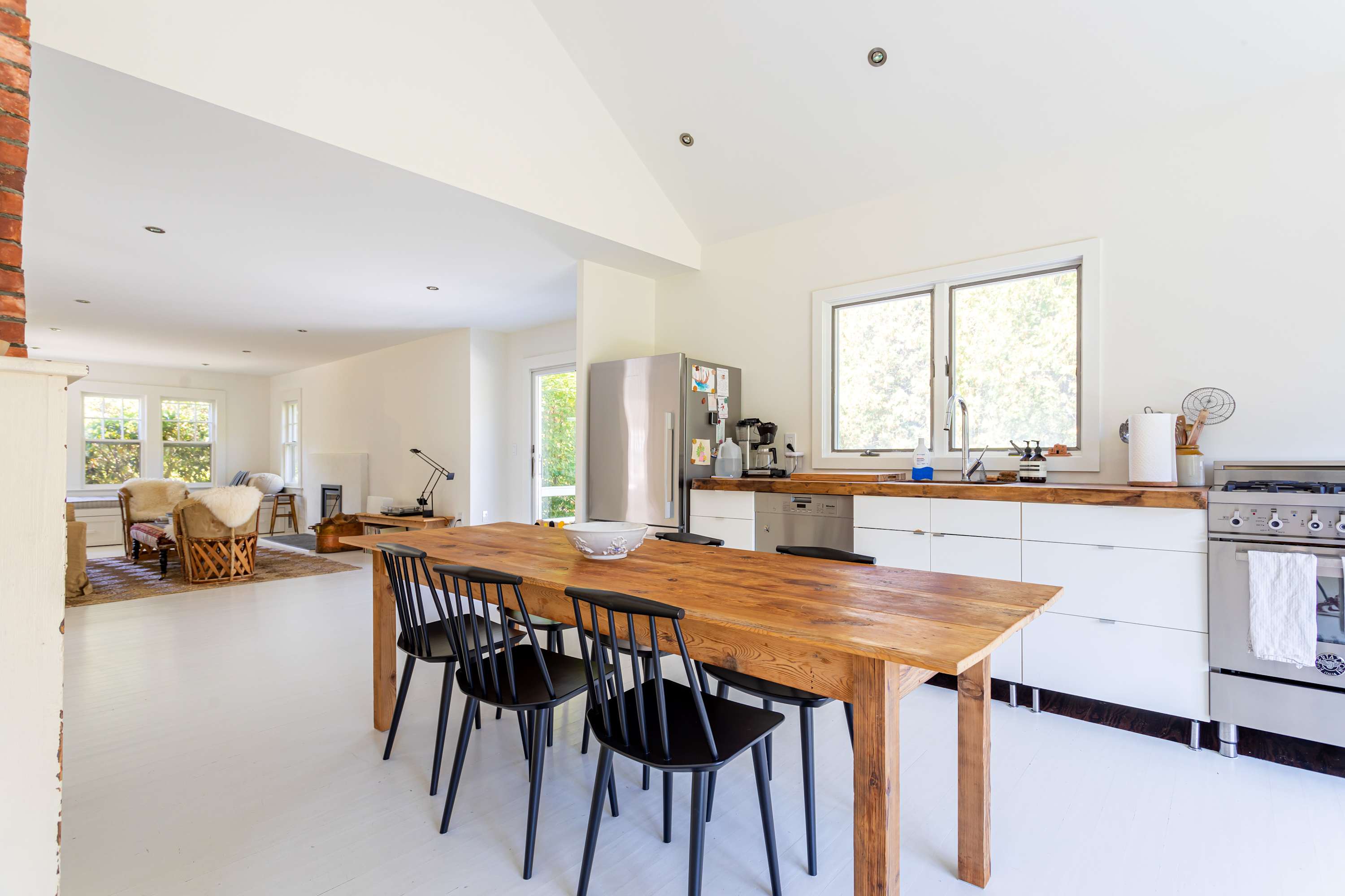 A bright, open-plan kitchen and dining space featuring a rustic wooden dining table with black chairs. White cabinetry and wooden countertops line the wall, with a stainless steel refrigerator and stove visible, all set on white floors.