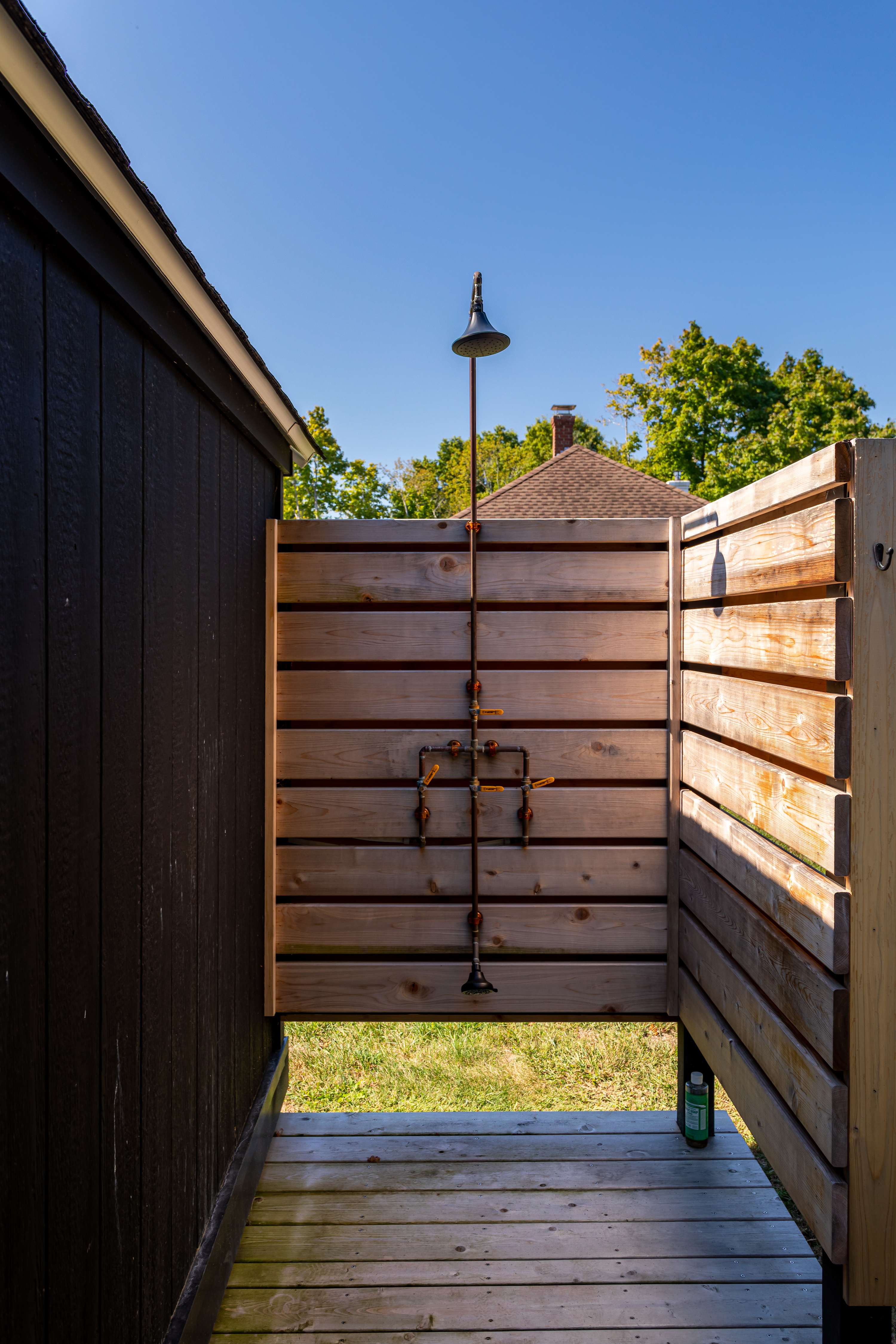 Rustic outdoor shower enclosed by natural wooden slatted walls, featuring exposed copper piping and a classic shower head, set on a wooden deck with grass visible beyond.