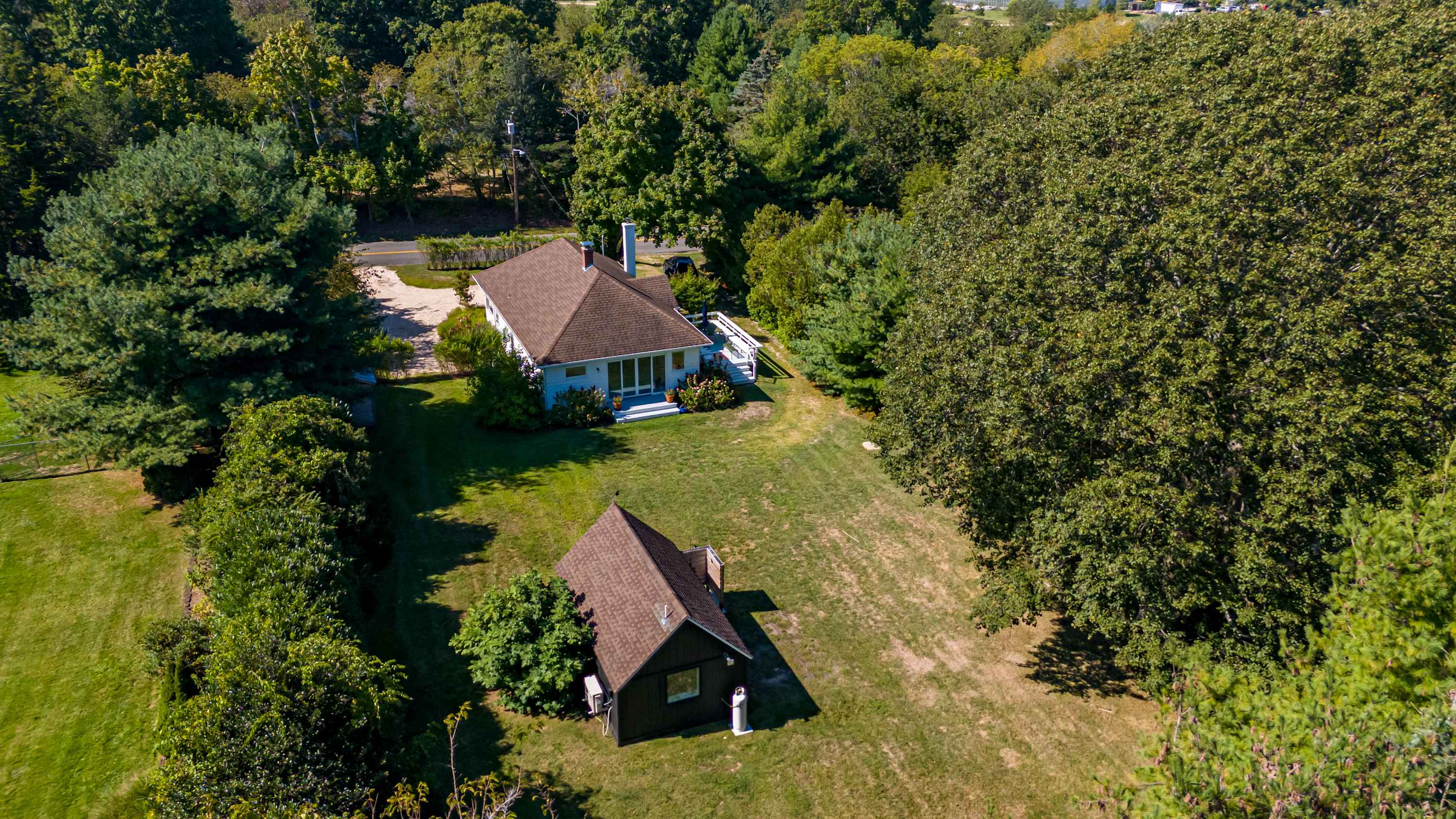 Aerial view showcasing the main house with a light-colored exterior and a brown roof, alongside a smaller dark-colored cottage, both nestled within a large green yard surrounded by lush trees.