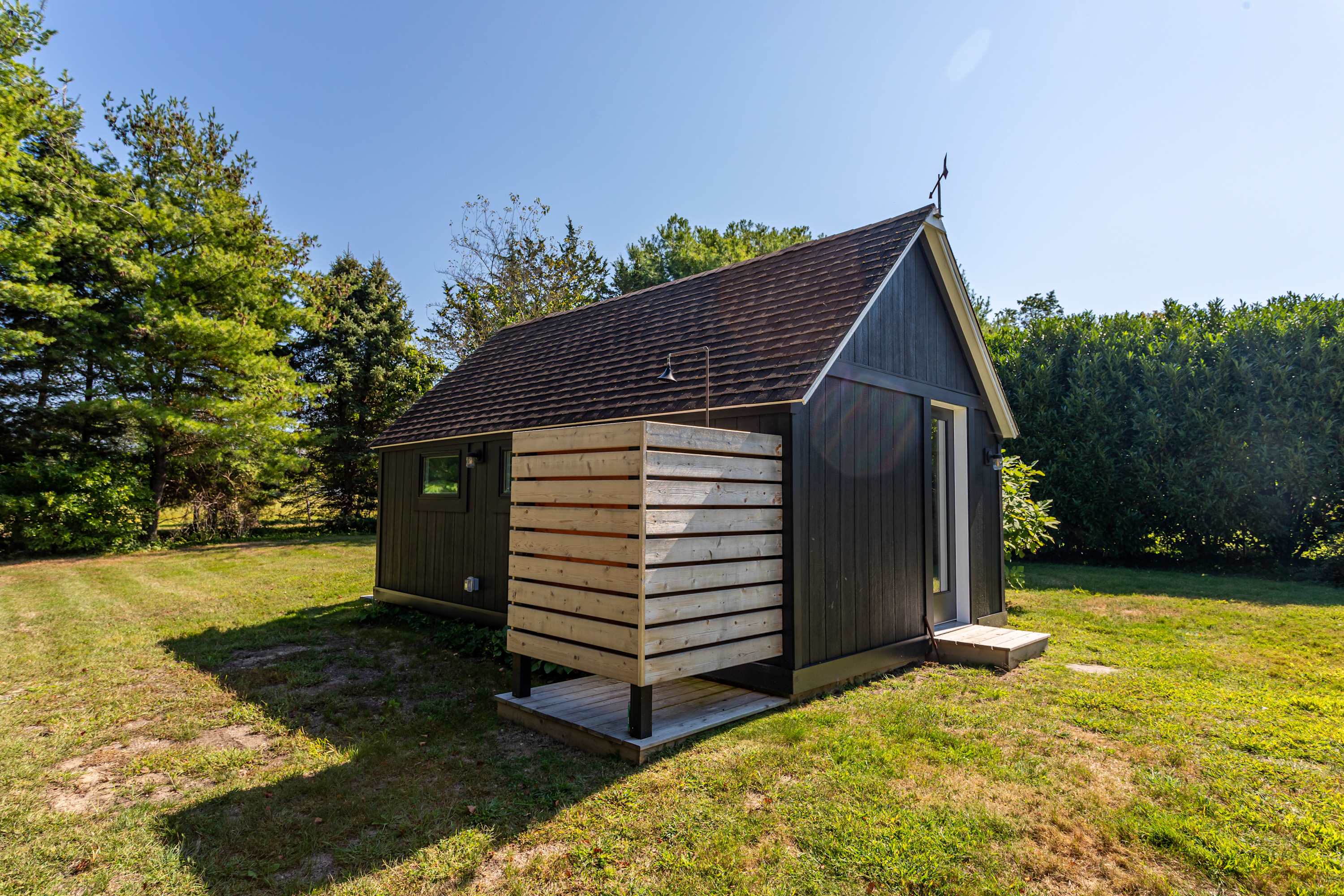 Dark-colored small cottage or shed with a brown roof, featuring a slatted wooden outdoor shower enclosure on one side, set in a grassy backyard with trees.