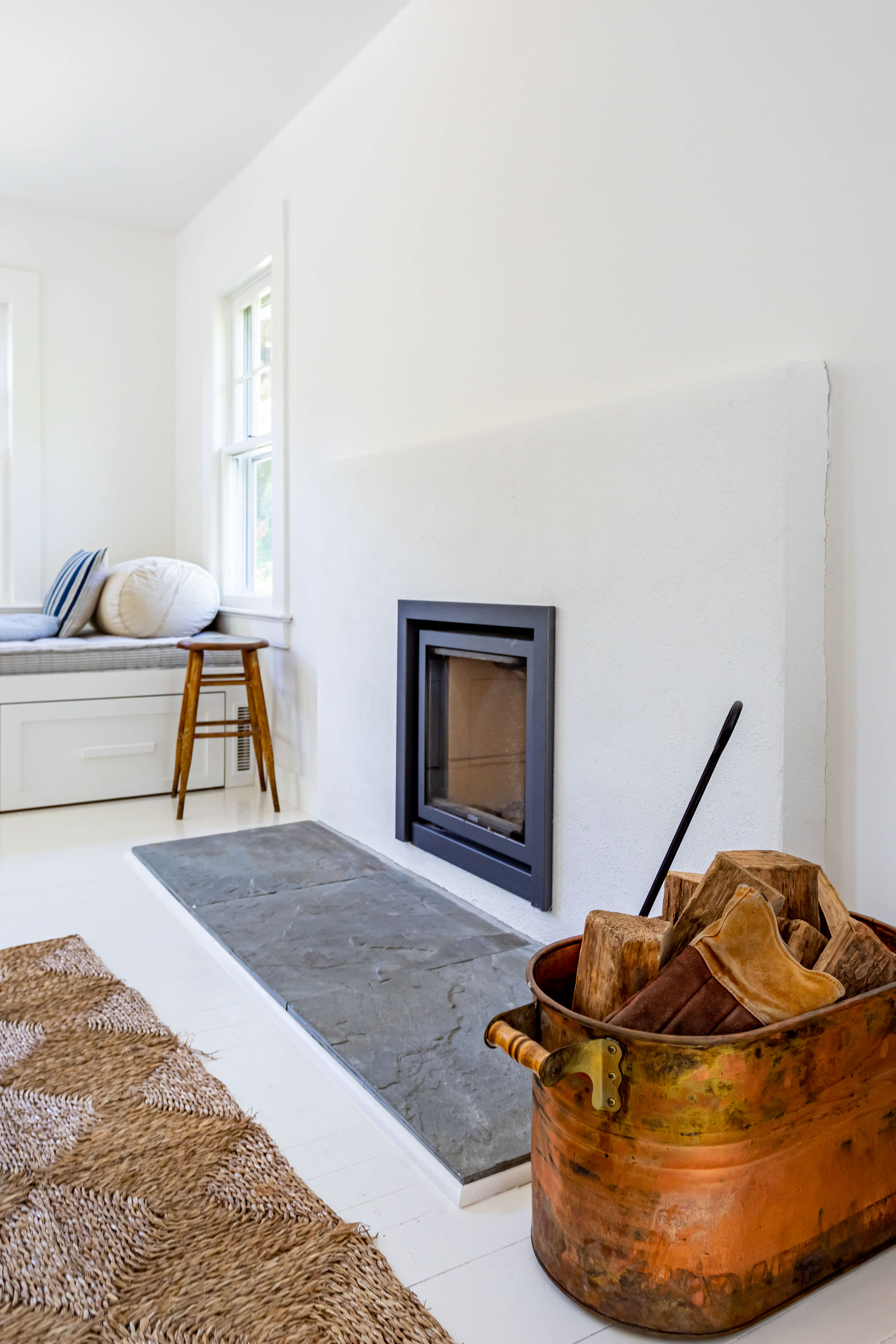Modern fireplace insert set into a minimalist white plastered wall, with a slate hearth and a copper bucket filled with firewood beside it, in a bright room with white floors.