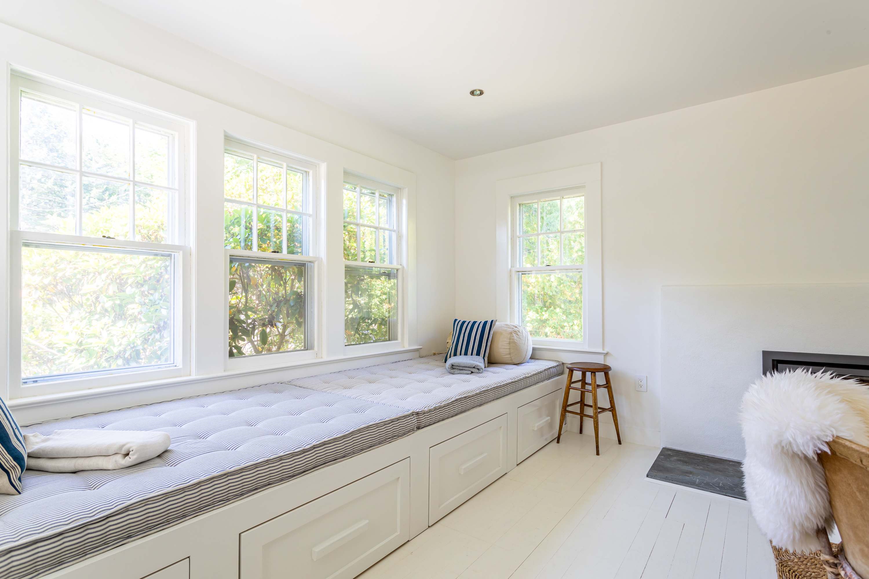 Bright window seat with a striped mattress and cushions, featuring built-in storage drawers underneath, offering views of the surrounding greenery through multiple windows.