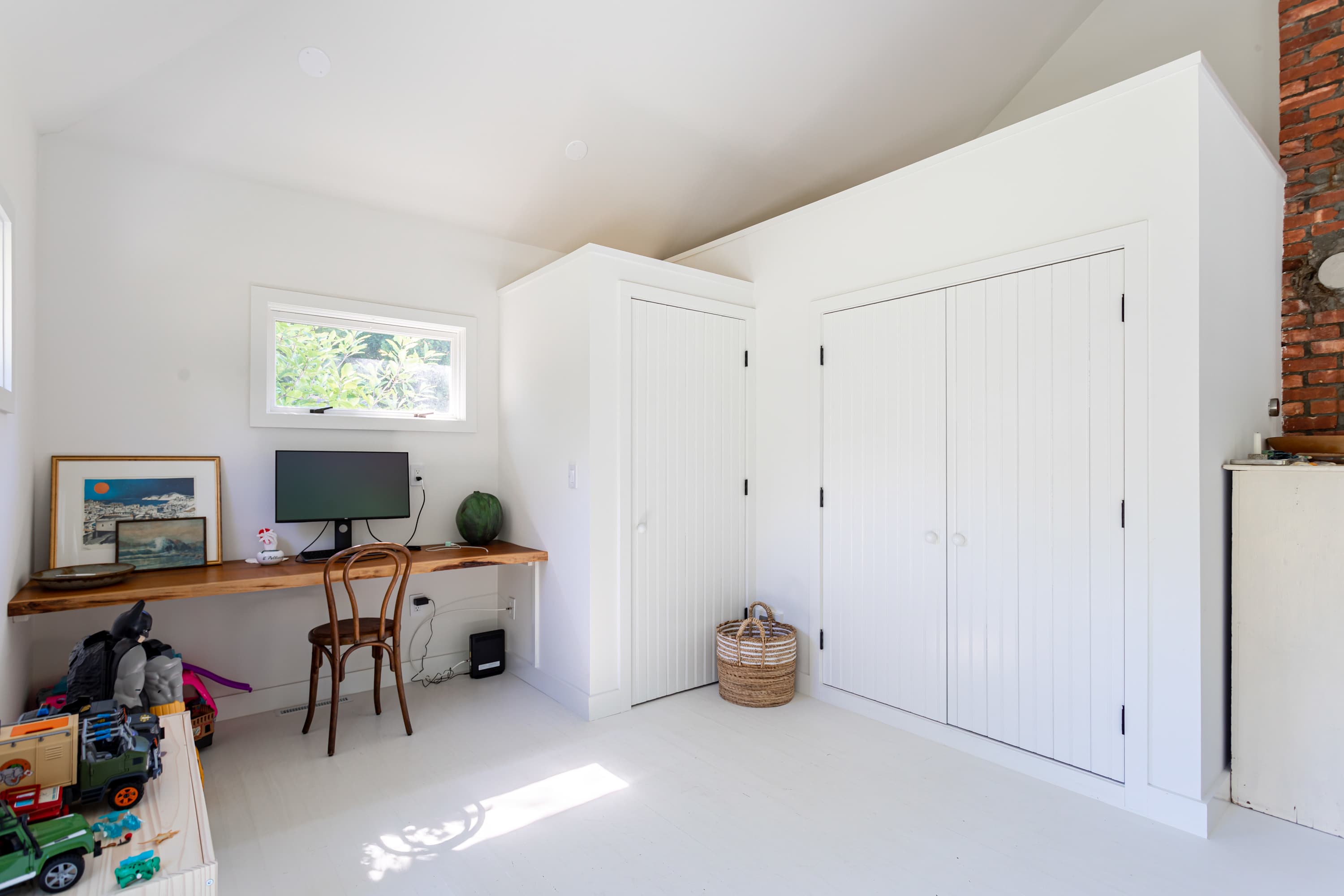 A bright home office space with white walls and floors, featuring a long wooden desk with a computer monitor and a simple wooden chair. Two white paneled closets are built into the wall, and a window provides natural light.