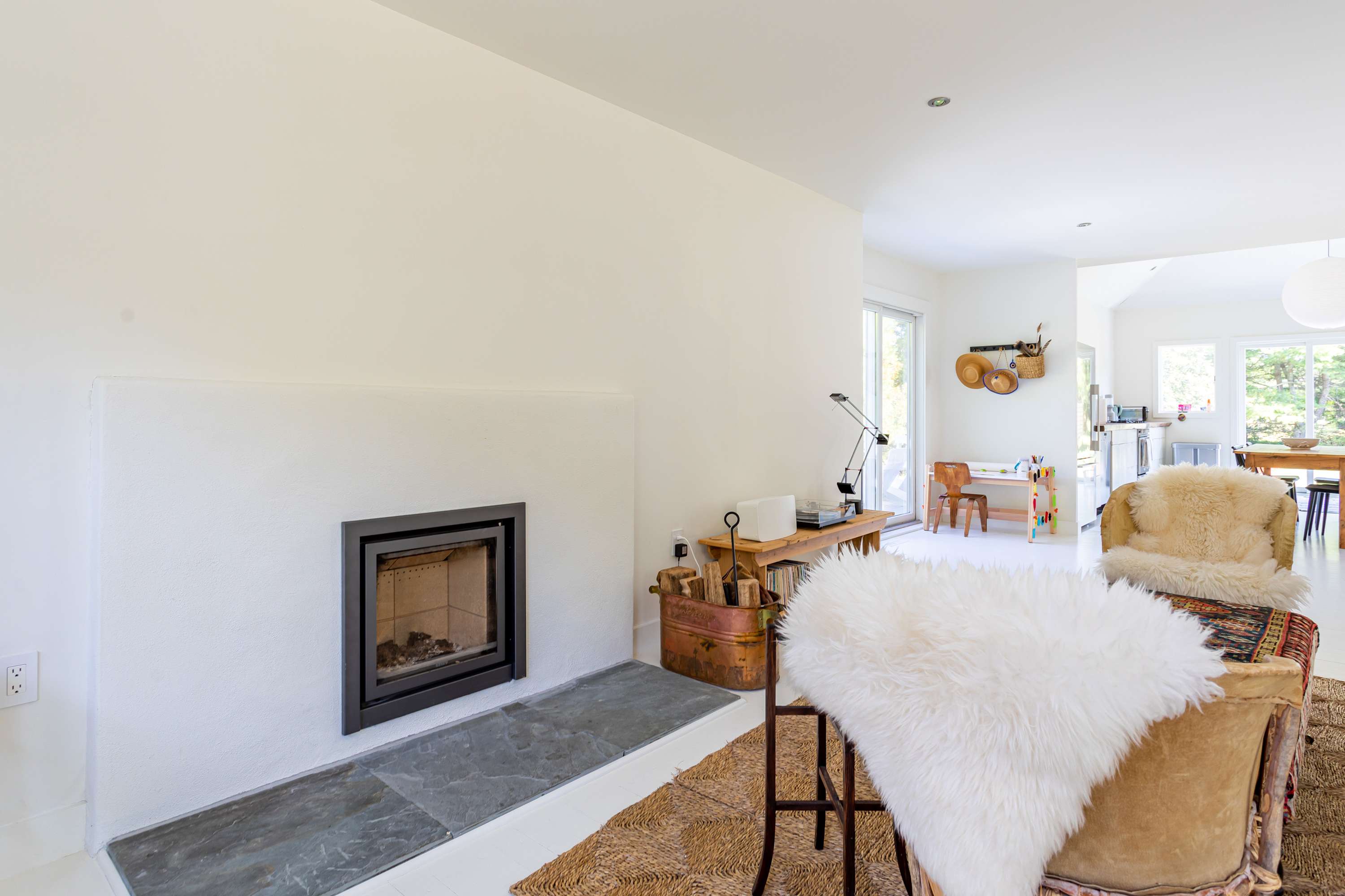 A cozy living room features a modern black fireplace insert set into a white wall, with a slate hearth and a rustic wooden bench holding firewood. A white faux fur throw drapes over a chair, and a woven rug covers the floor.