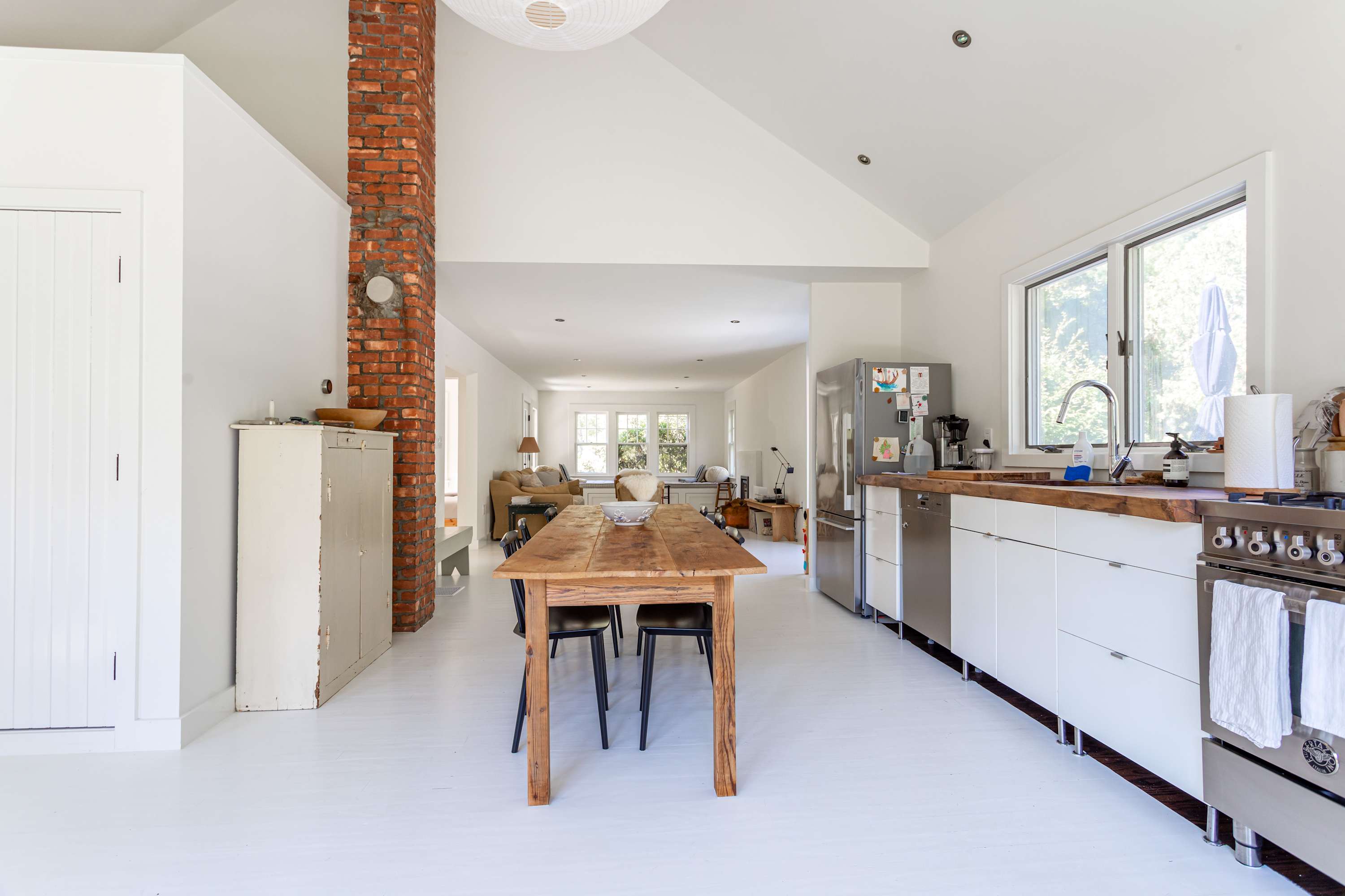 An expansive kitchen and dining area with white floors and vaulted ceilings, featuring a long wooden dining table and black chairs. The kitchen has white cabinets, wooden countertops, and stainless steel appliances, with a prominent brick column.