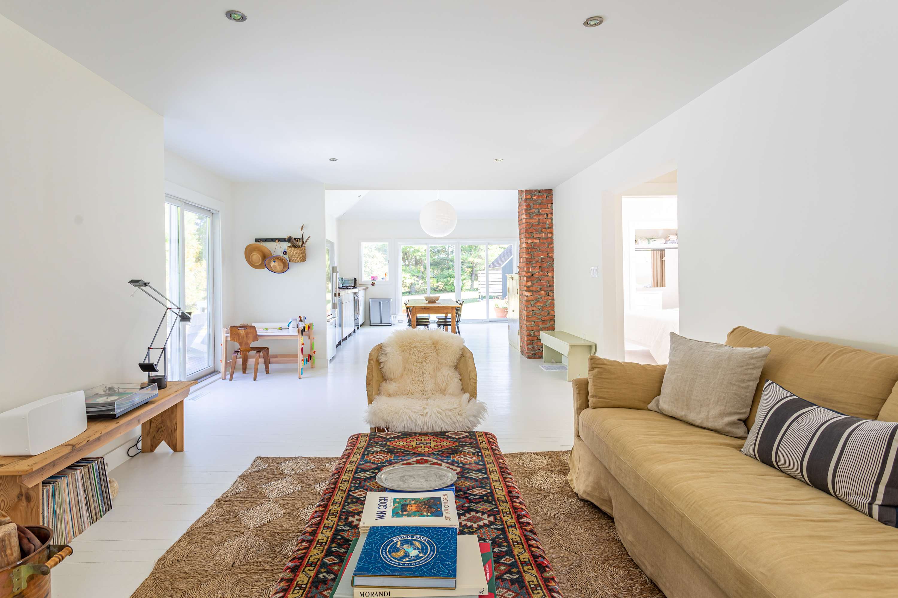 An open-concept living room with a mustard yellow sofa and a colorful patterned rug, leading into a bright kitchen and dining area. A white faux fur chair and a wooden coffee table are central, with white walls and floors throughout.