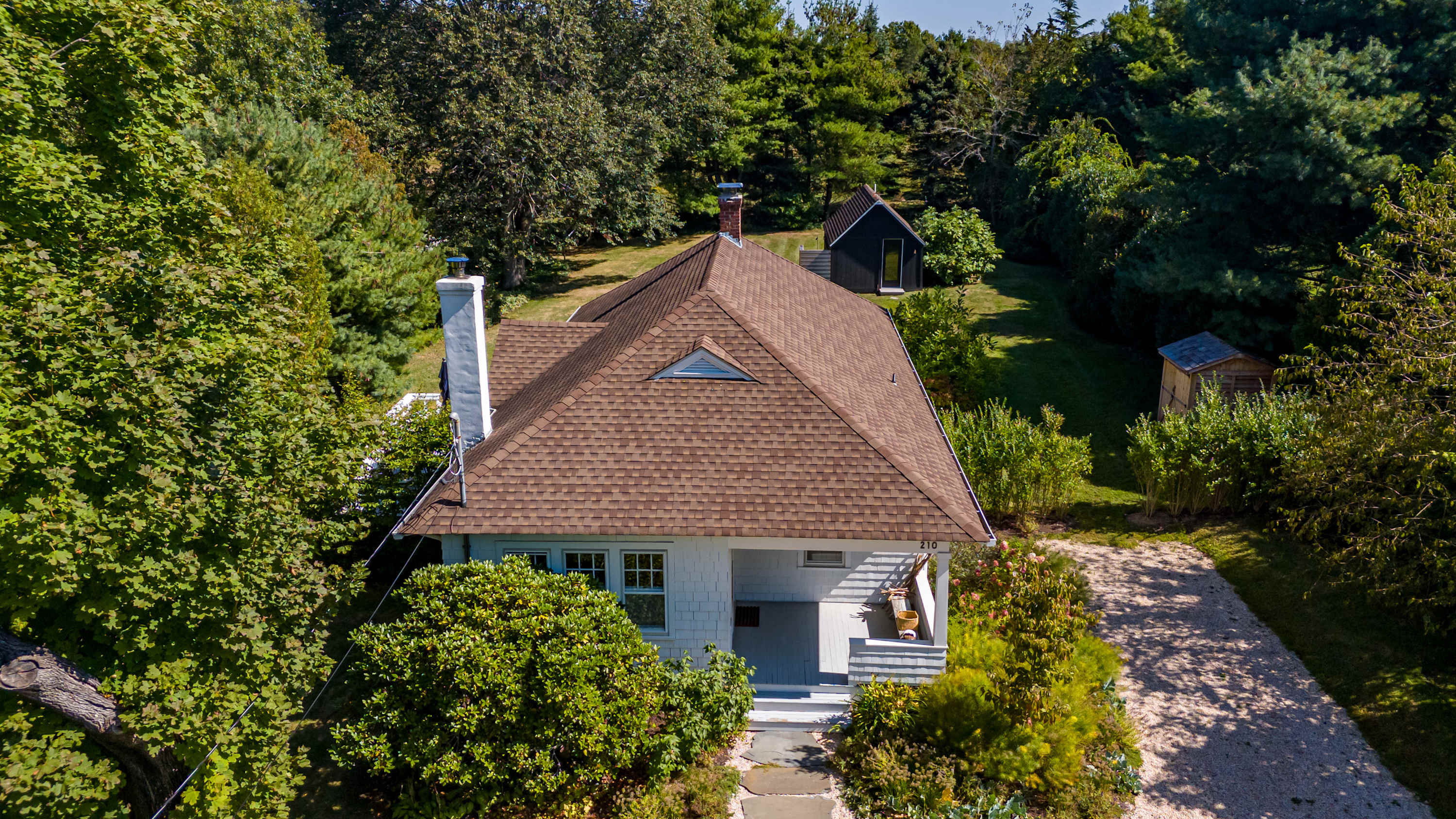 An aerial view of a charming cottage with a brown shingled roof and white siding, nestled among dense green trees, featuring a gravel driveway and a small black outbuilding in the background.