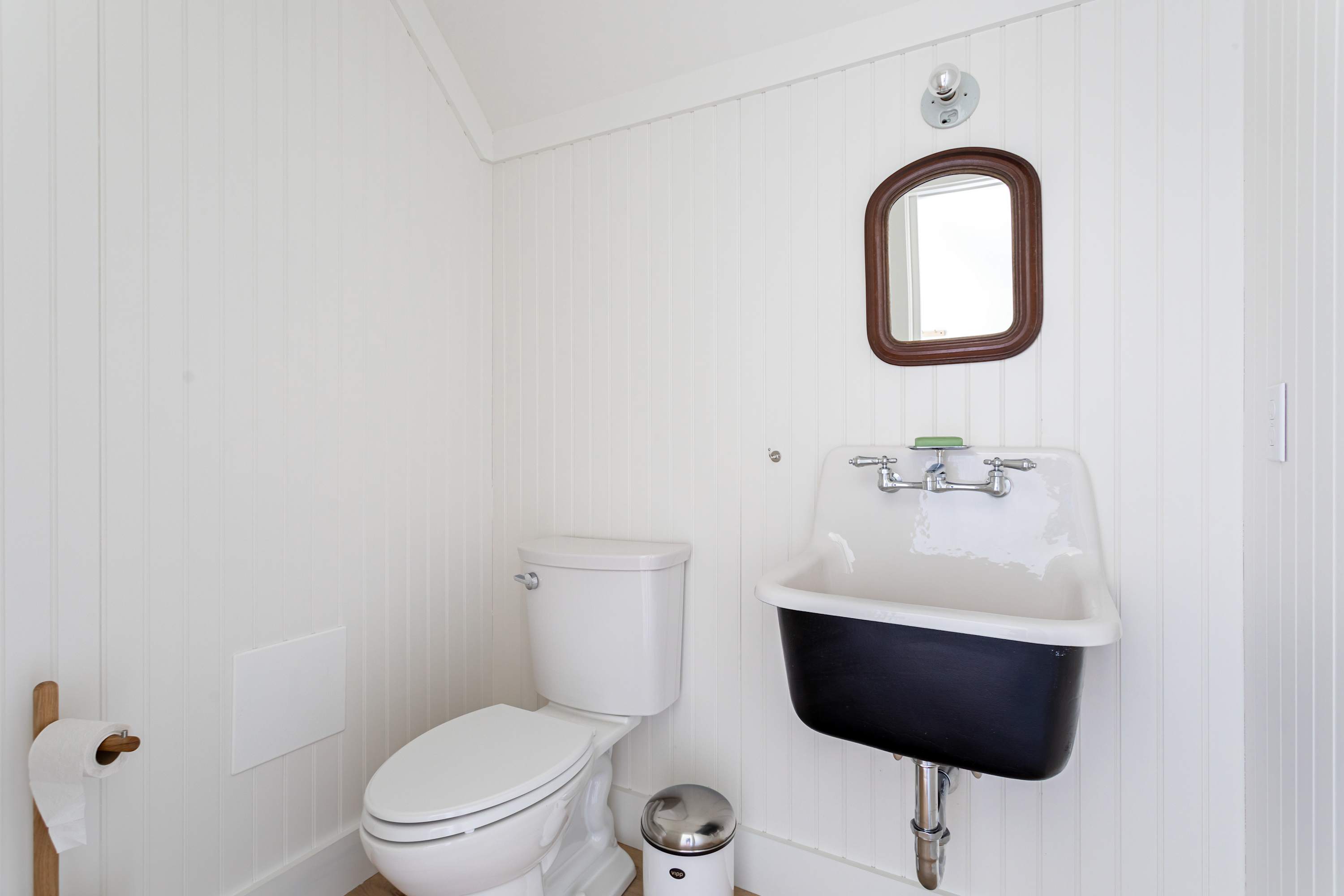 Compact half-bathroom with white beadboard walls, featuring a white toilet, a vintage-style black and white sink with chrome fixtures, and a small wooden-framed mirror.