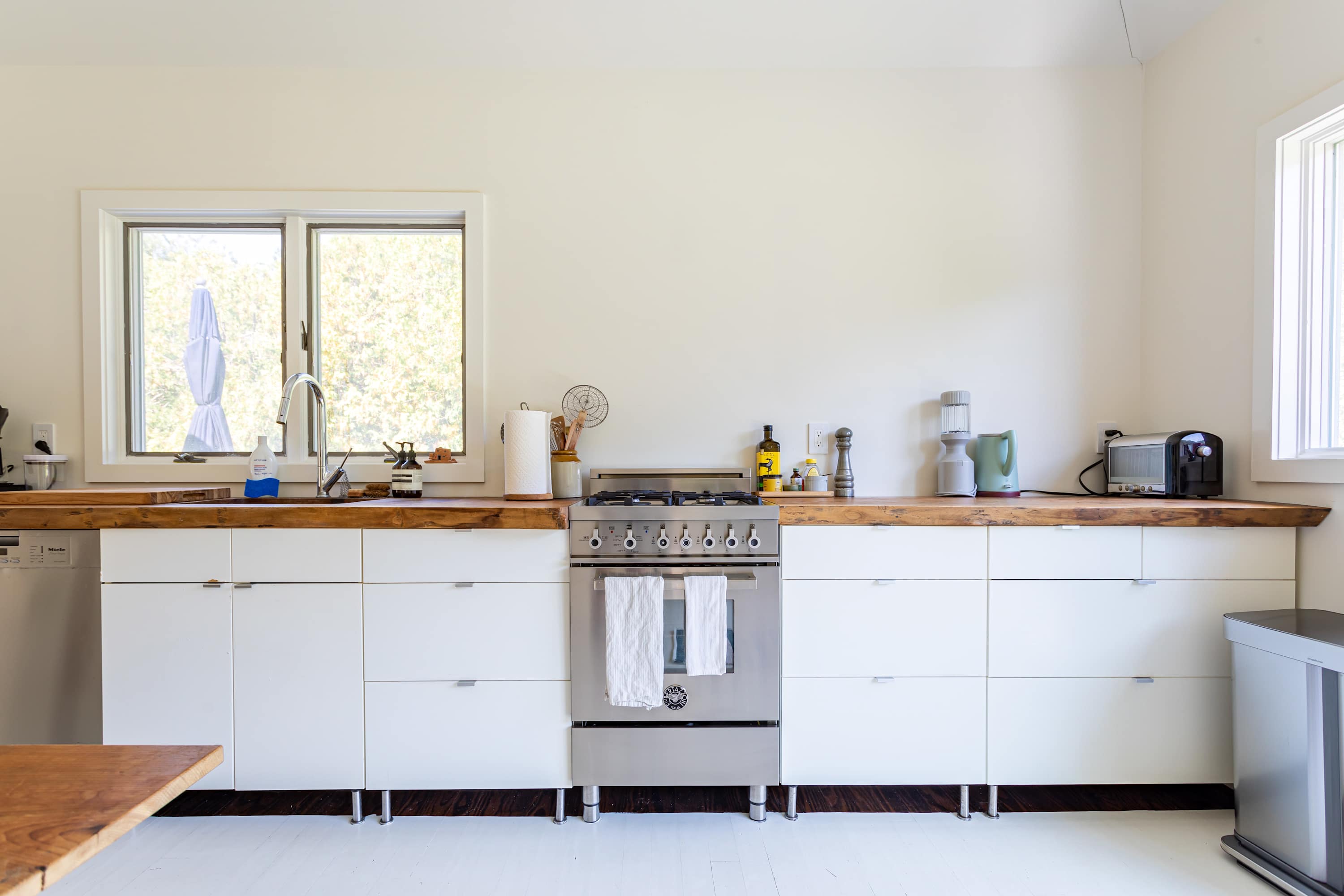 A bright kitchen counter with light wooden countertops, white base cabinets, and a stainless steel range with a double oven. A window above the sink provides natural light, and various kitchen tools are neatly arranged.