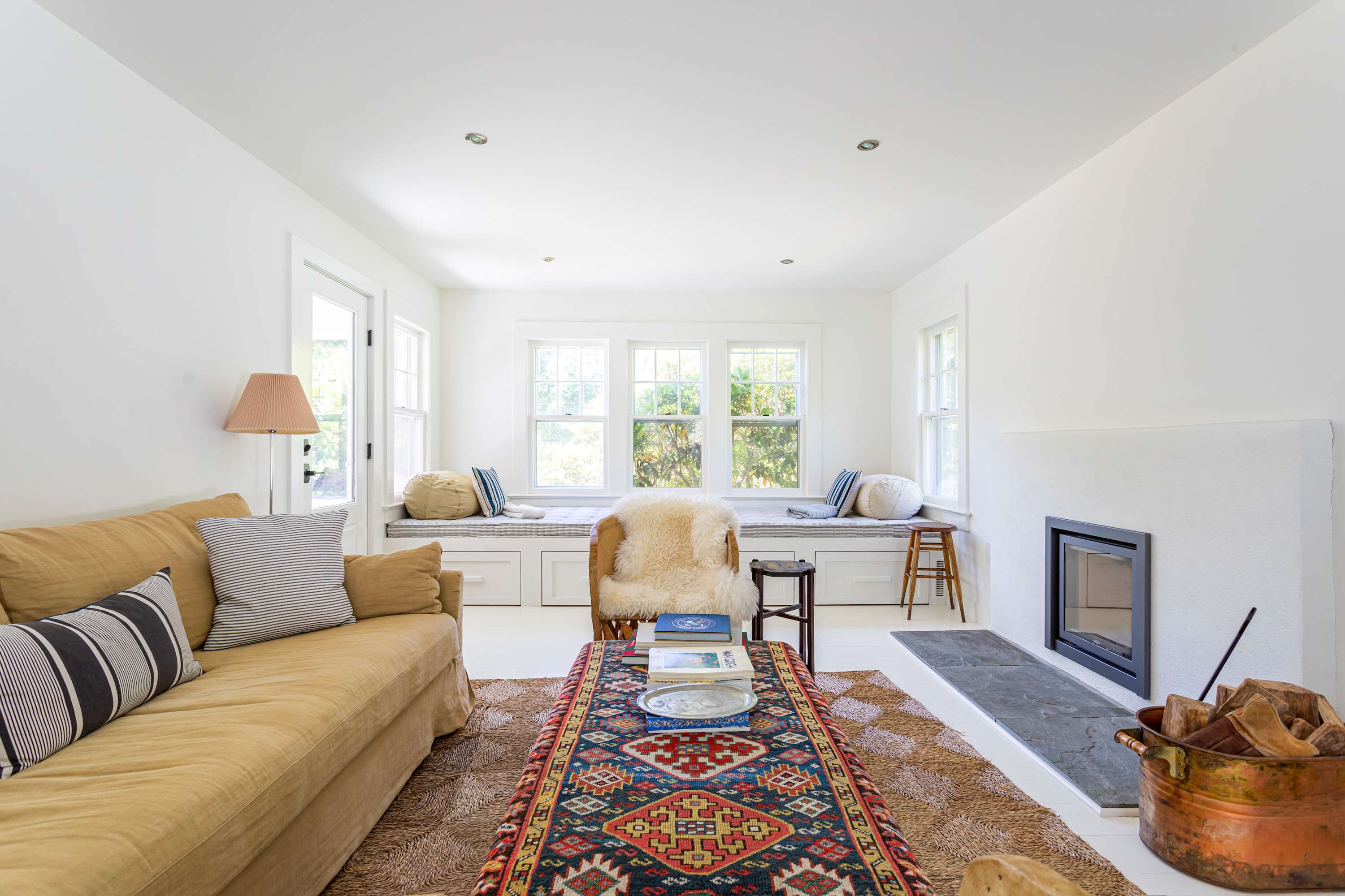 A bright living room with a mustard yellow sofa, a colorful patterned rug over a natural fiber rug, and a coffee table stacked with books. Window seats with cushions line the far wall, offering a view of the outdoors.