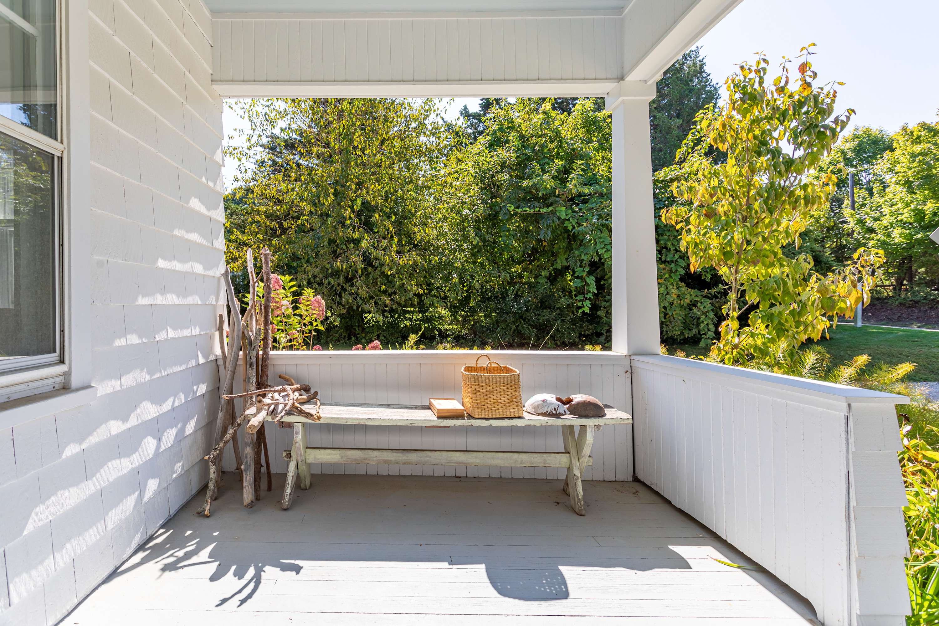 Covered front porch with white shiplap walls, a rustic wooden bench adorned with a woven basket and decorative items, offering a serene view of the lush green yard.