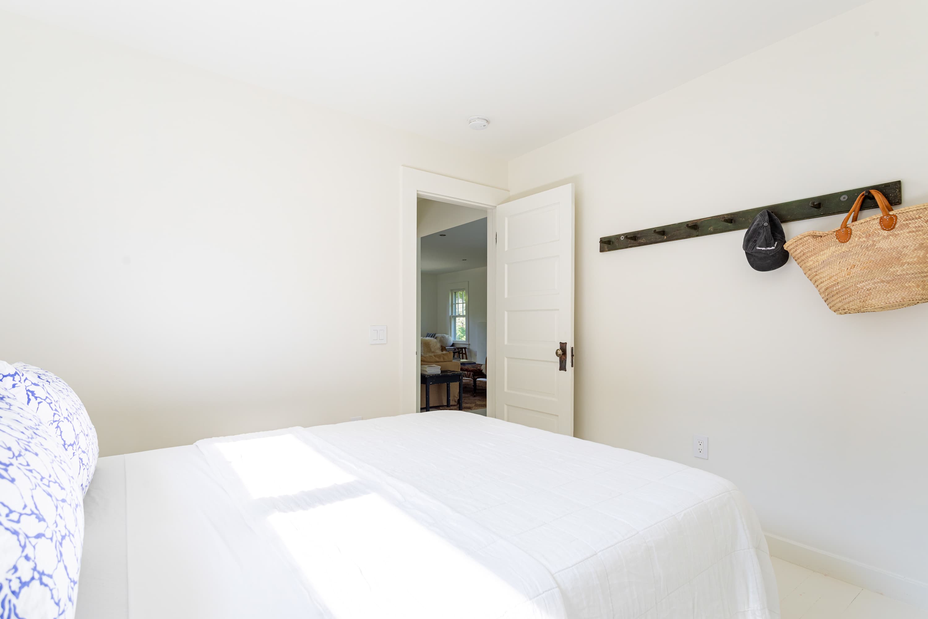 Bright bedroom with white walls and bedding, featuring a wooden coat rack with a black hat and straw bag, and a doorway leading to a living area.