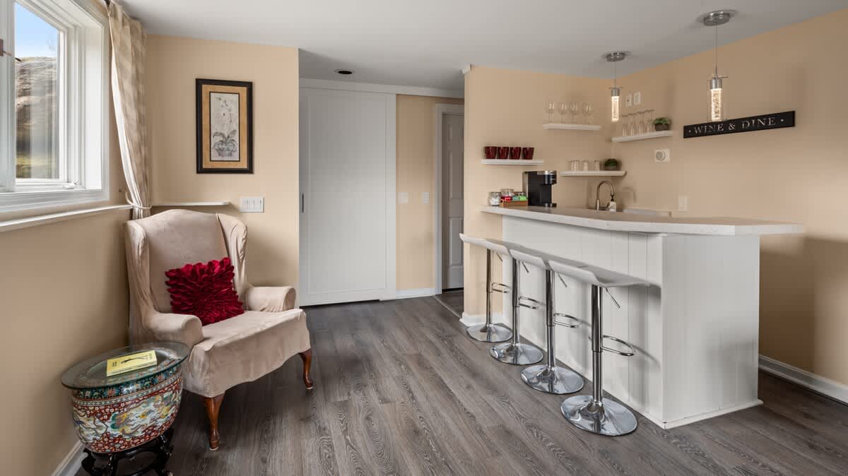 A bright corner of the living room featuring a white wet bar with three chrome bar stools, a sink, and open shelving with glassware. A light-colored armchair with a red pillow sits by a window.
