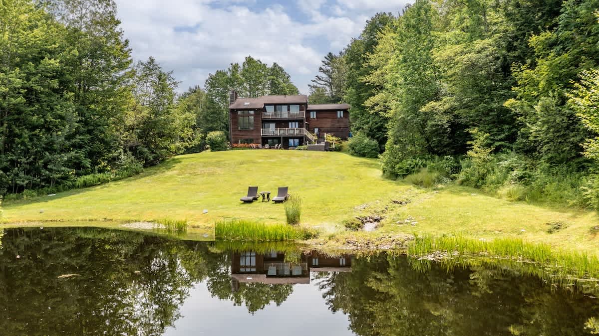 A large, multi-story wooden house with multiple decks overlooks a manicured green lawn sloping down to a reflective pond, surrounded by dense forest under a partly cloudy sky.