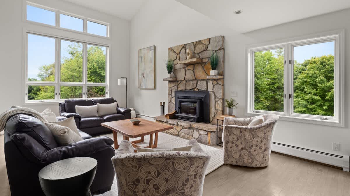 This bright living room boasts a tall stone fireplace, comfortable seating including leather sofas and patterned armchairs, and expansive windows that provide abundant natural light and views of the lush outdoor landscape.