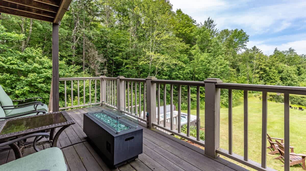 A wooden deck features outdoor seating and a rectangular fire pit with glass rocks, overlooking a lush green lawn and dense forest with a hot tub visible in the distance.