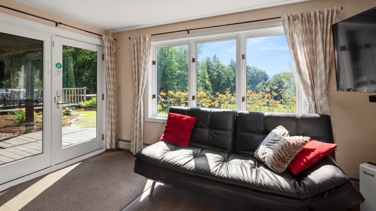 A bright living space with a black futon sofa, red and patterned throw pillows, and large windows offering a lush green outdoor view with yellow flowers. A glass door leads to an outdoor deck.