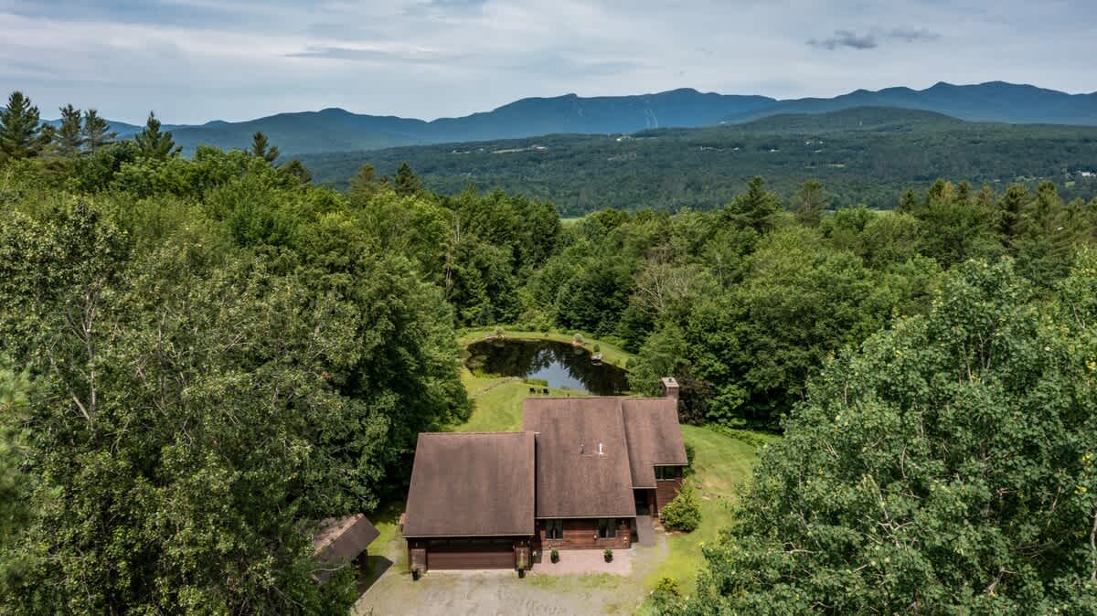 An aerial view reveals a large house with a dark brown roof nestled among lush green trees, featuring a small pond in the backyard and distant mountains under a cloudy sky.