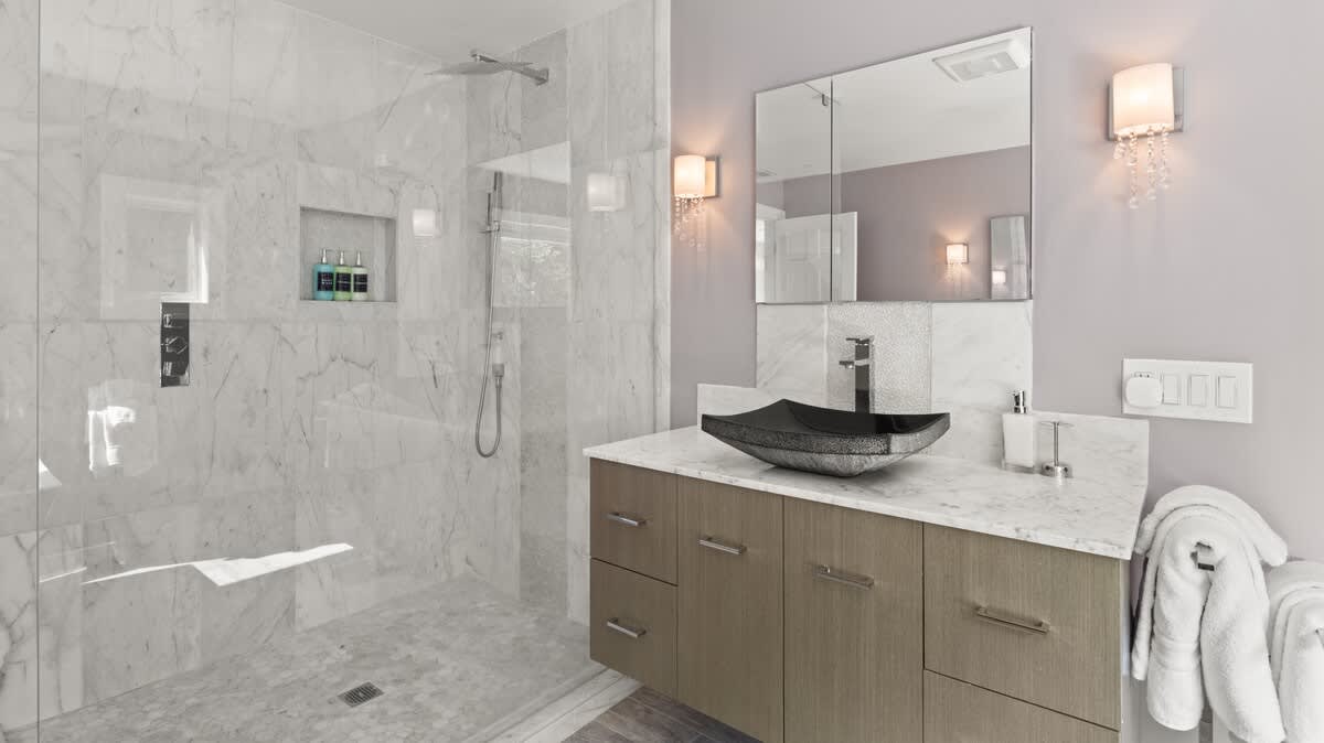 Contemporary bathroom featuring a spacious walk-in shower with white marble-patterned tiles and a glass enclosure. A light wood vanity with a black vessel sink and marble countertop is visible, along with wall sconces and a large mirror.
