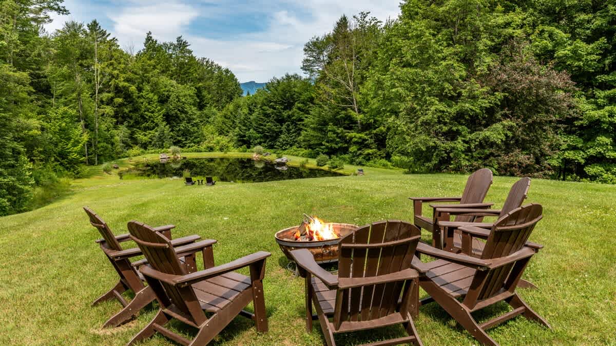 Outdoor fire pit area with several dark wooden Adirondack chairs arranged around a burning fire pit on a green lawn. A tranquil pond is visible in the background, surrounded by dense forest and distant mountains.
