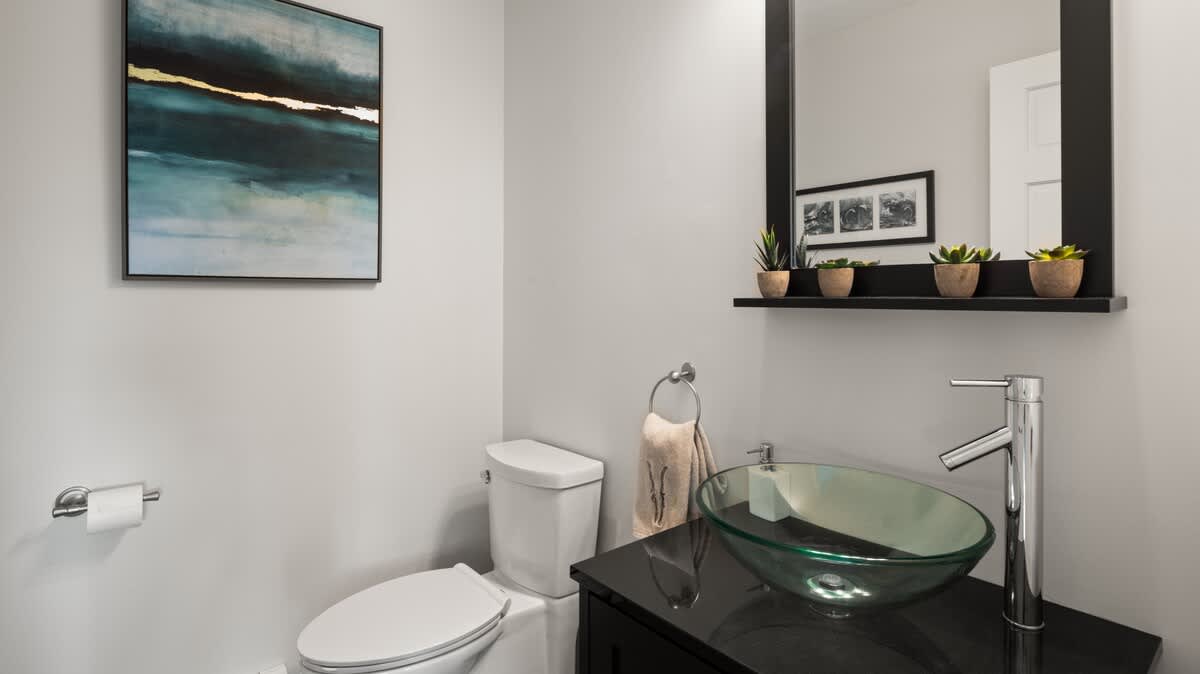 Compact powder room with light gray walls, featuring a white toilet and a black vanity with a green glass vessel sink. A black framed mirror with a shelf holding small potted plants hangs above the sink, complemented by modern artwork.
