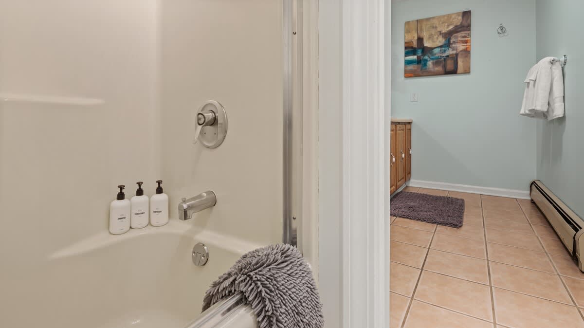 A view of a bathtub with a shower head, featuring three labeled bottles of bath products. The room has light blue walls and a tiled floor, visible through the doorway.
