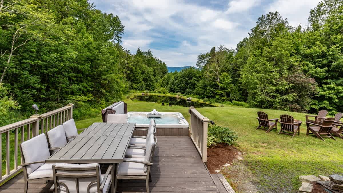 An outdoor wooden deck features a dining table with chairs and a hot tub, overlooking a vast green lawn, a pond, and a dense forest with distant mountains under a partly cloudy sky.