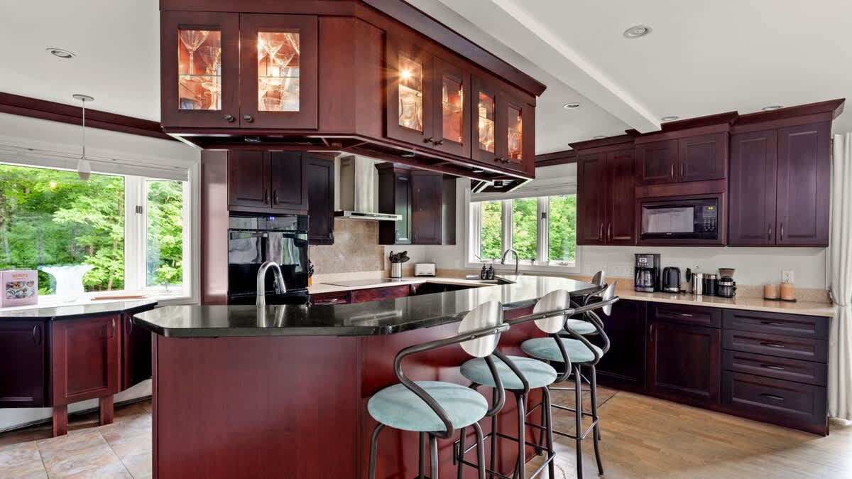 A modern kitchen features dark cherry wood cabinetry, black granite countertops, stainless steel appliances, and a large island with bar seating, all illuminated by under-cabinet lighting and natural light from large windows.