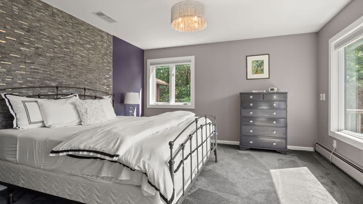 A modern bedroom features a metal-frame queen bed with white bedding, a textured gray accent wall, a gray dresser, and large windows offering views of the surrounding trees.